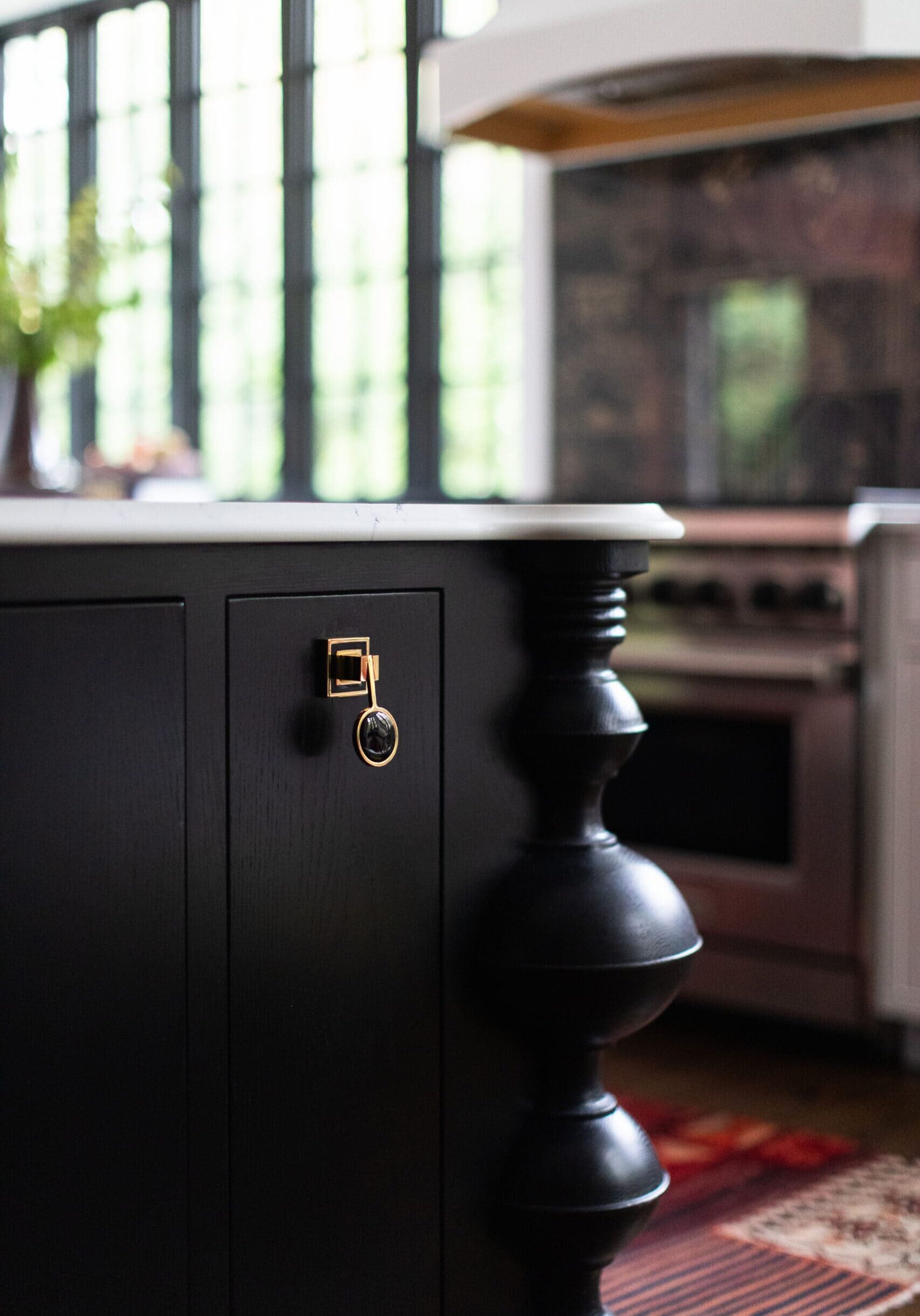 Close-up of a black kitchen island with a marble countertop, featuring ornate turned wood detailing and a gold drawer pull; a stove and large windows are visible in the background.