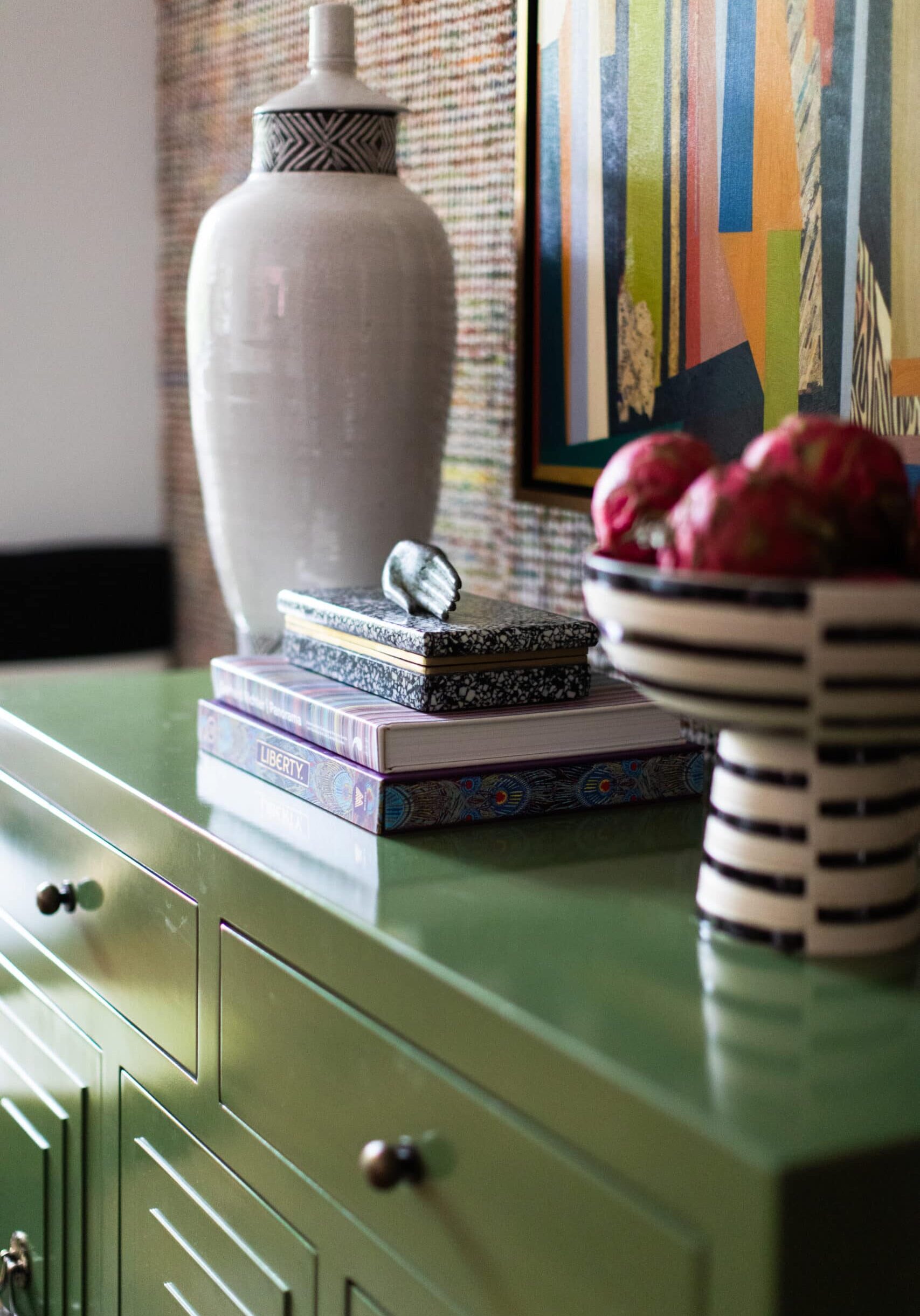A white ceramic vase, two books, a decorative box, and a striped bowl of red fruit are arranged on a glossy green cabinet beneath a colorful abstract painting.
