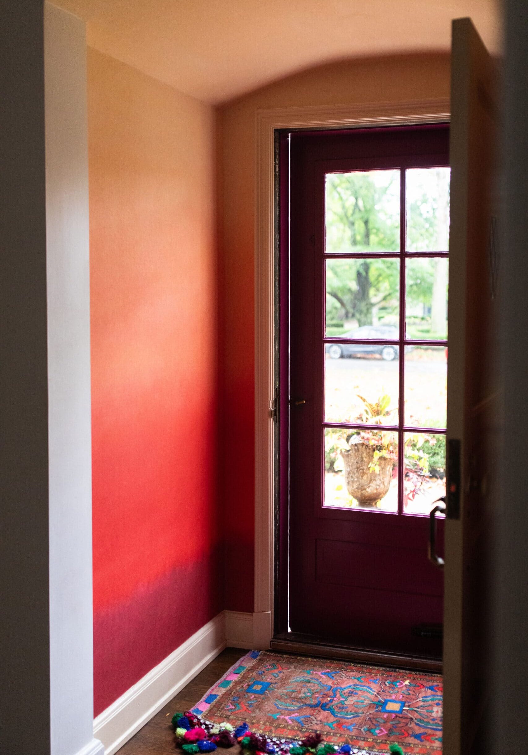 A front door with glass panels lets in daylight. A colorful rug and a string of pom-poms are on the floor by the entrance. Trees and a car are visible outside.