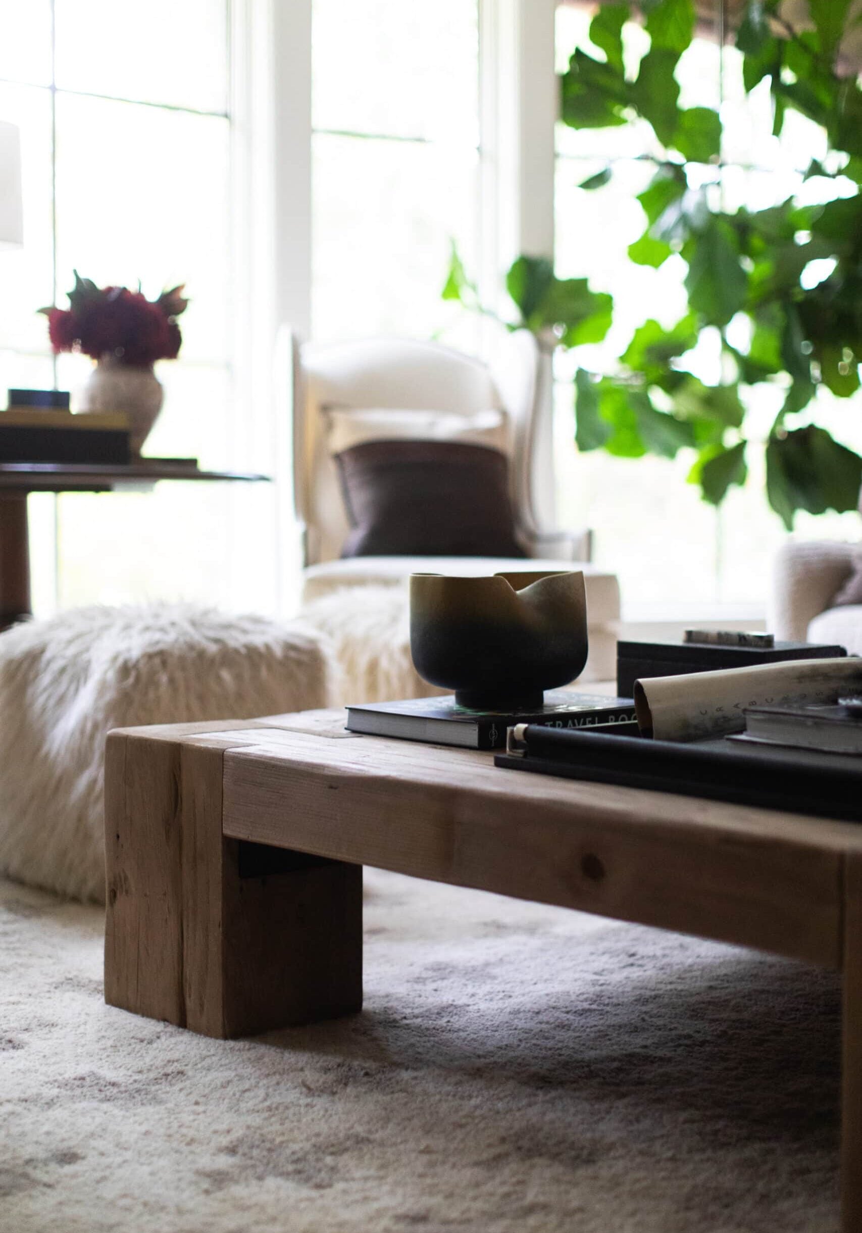 A low wooden coffee table with books and a candle sits on a light carpet in a cozy living room with large windows, a chair, and green plants in the background.