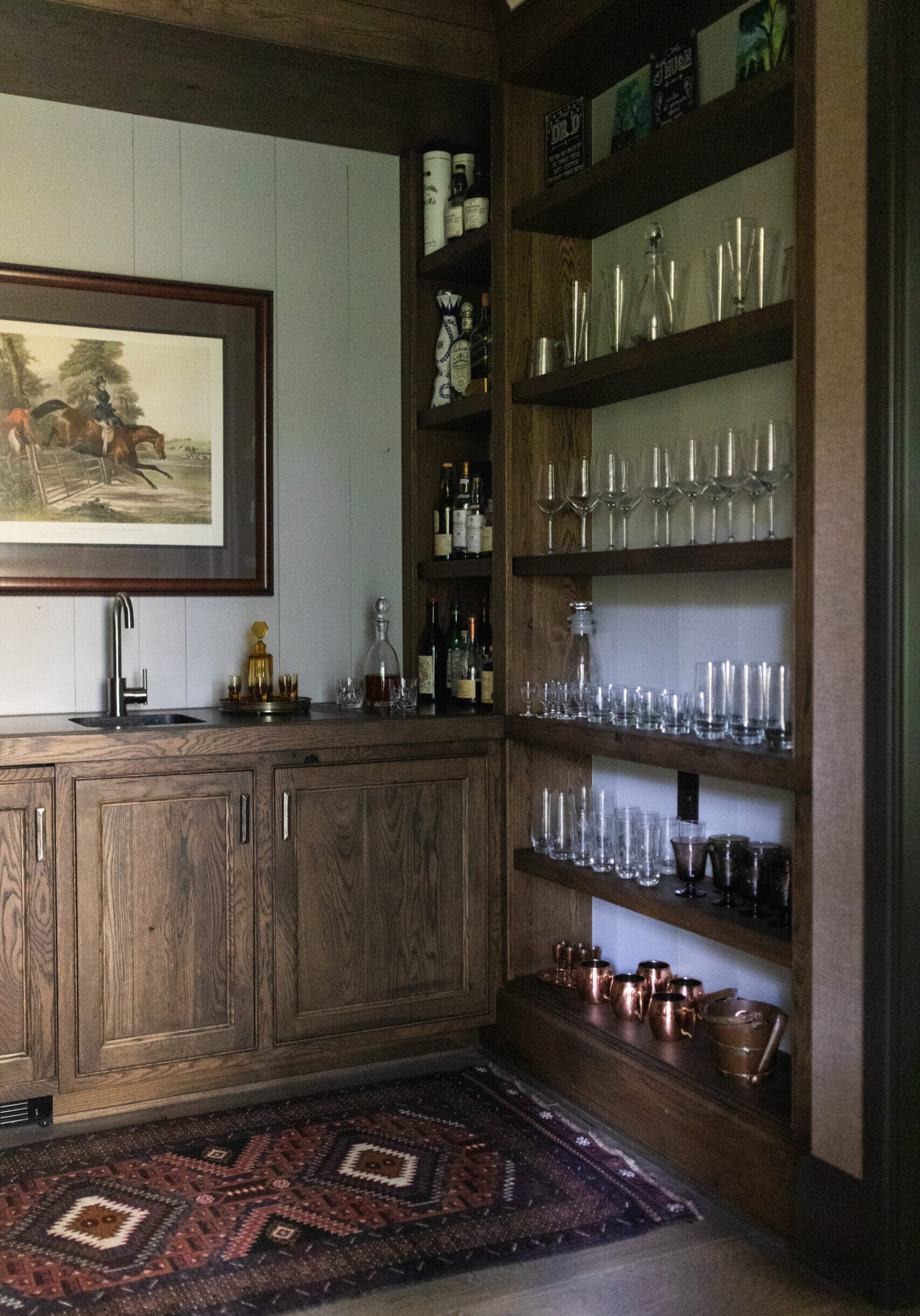Home bar area with wooden cabinets, a small sink, shelves of glassware, bottles, and copper cups, next to a large framed painting and patterned rug.