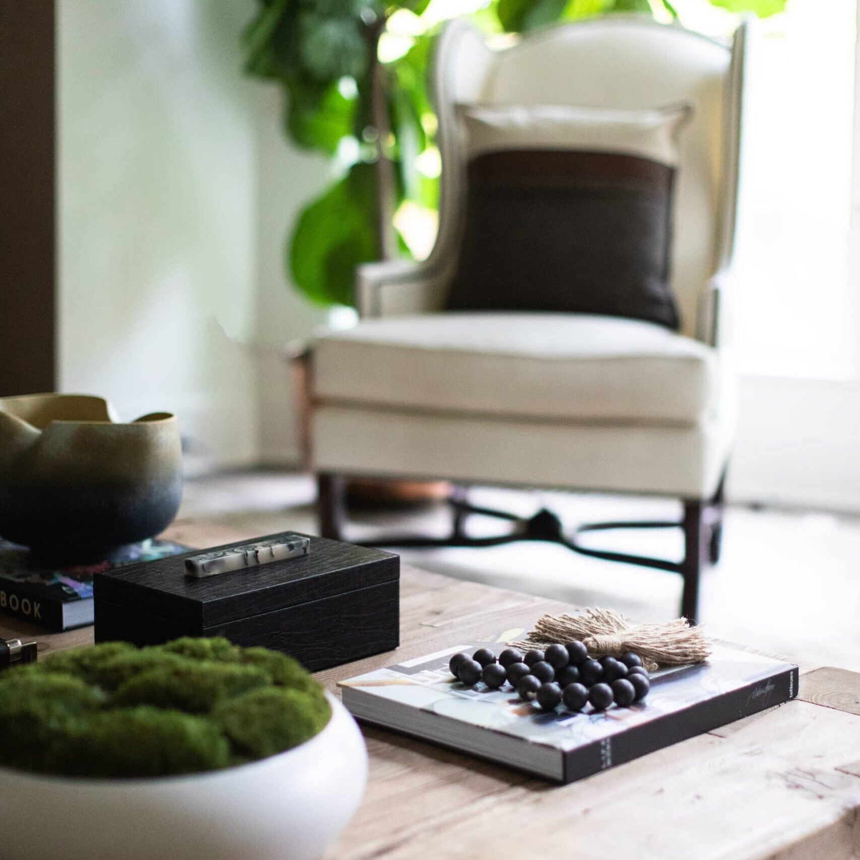 A wooden coffee table with decorative items in front of a white armchair, brown pillow, and large green plant by a bright window.