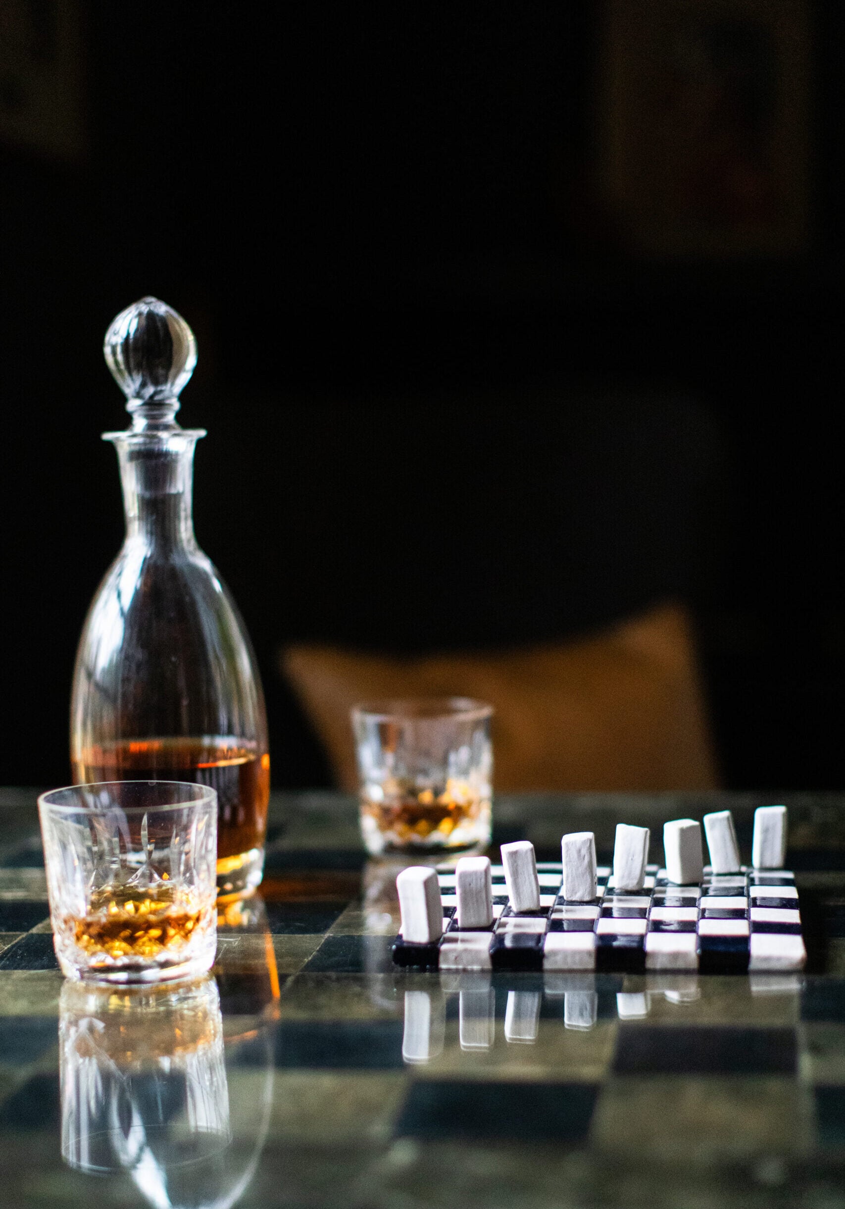 A glass decanter with amber liquid, two filled whiskey glasses, and a domino game set on a checkered table.