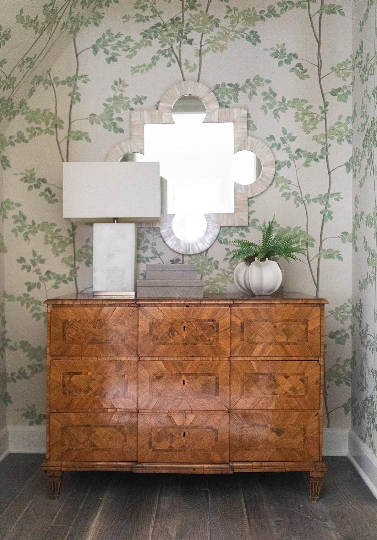 A wooden dresser with a geometric pattern is topped with a rectangular lamp and a white vase holding a green plant, set against wallpaper with a leafy vine design and a decorative mirror.