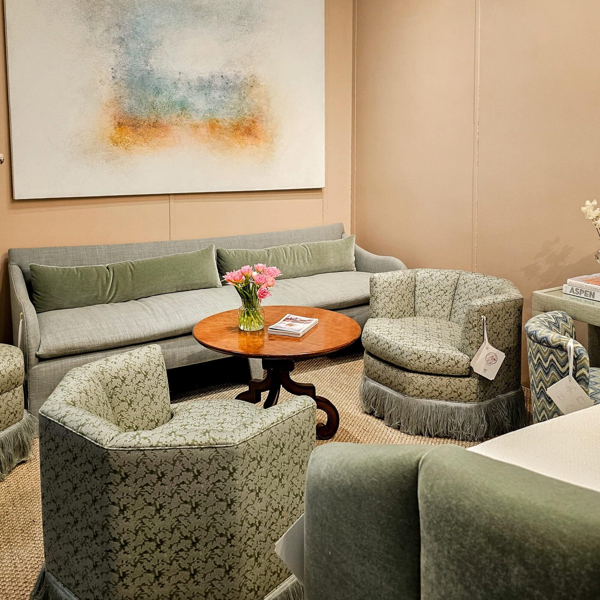 Sage green fringed accent chairs and a gray upholstered sofa surrounding a round walnut pedestal coffee table, spotted at High Point Market spring 2026.