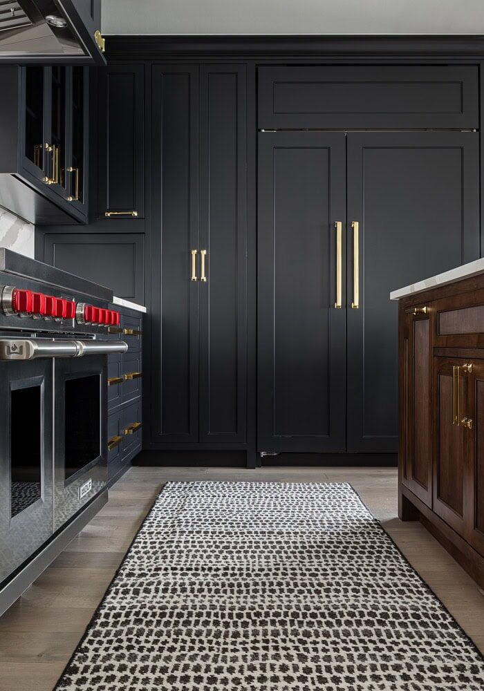 Modern kitchen with black cabinets, gold handles, stainless steel stove, dark wood island, marble backsplash, and a patterned runner rug on light wood floors.