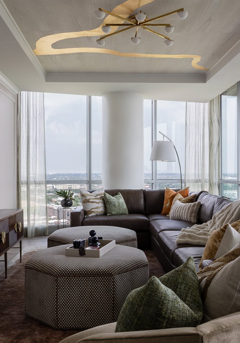 Modern living room with a large sectional sofa, multiple cushions, a geometric ottoman, floor lamp, and floor-to-ceiling windows overlooking a cityscape.