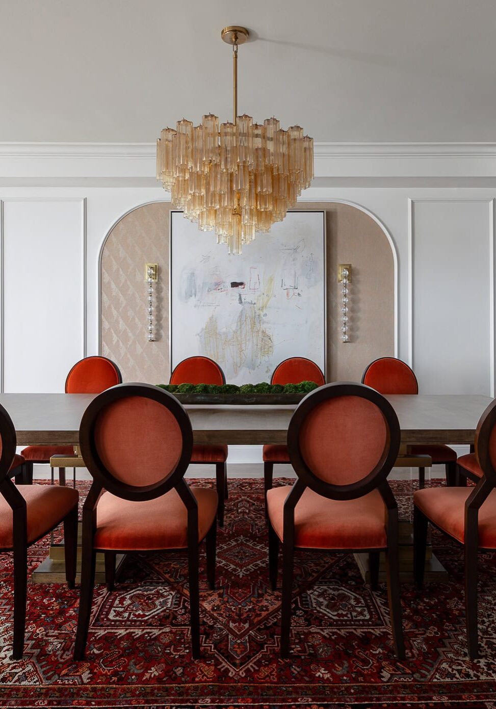A dining room with a modern chandelier, red upholstered chairs, a large dining table, abstract wall art, and a patterned red rug.