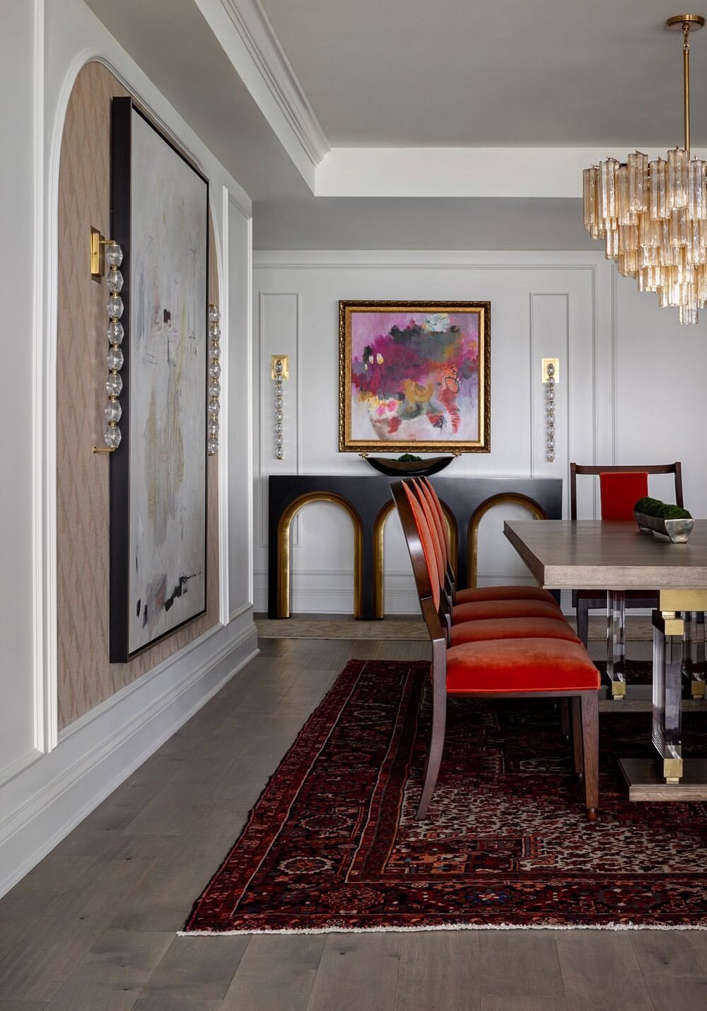 A modern dining room with a wooden table, red upholstered chairs, Persian rug, abstract wall art, and a gold-accented console under a framed painting.
