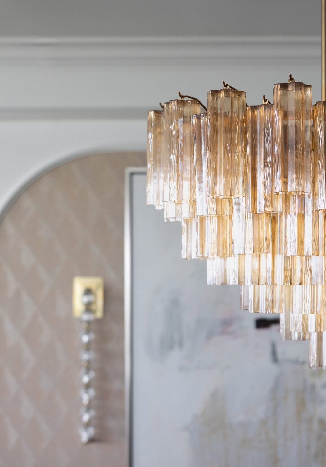 A close-up of a modern glass chandelier with vertical crystal rods, hanging in a room with light-colored walls and a blurred background.