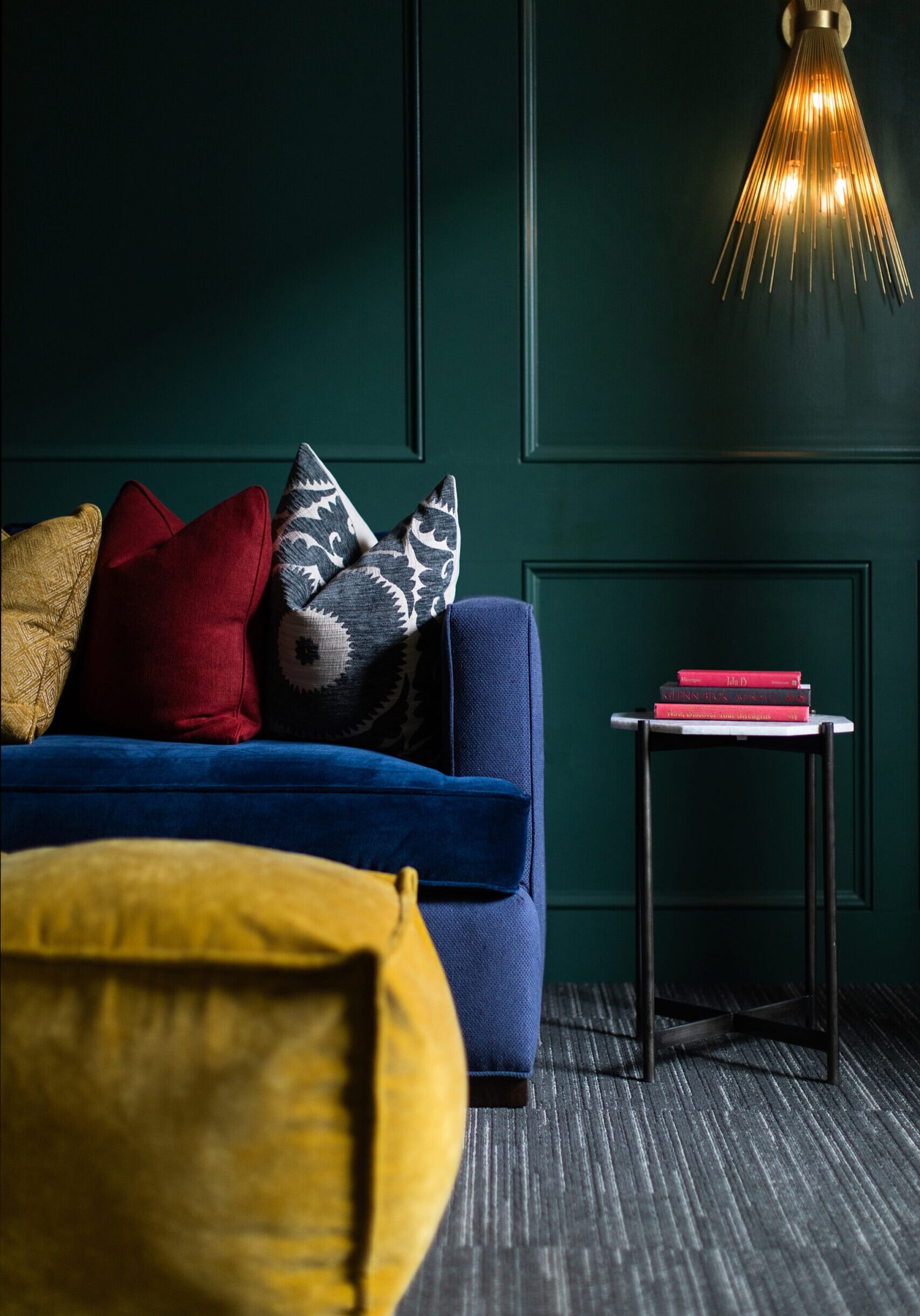 A modern living room with a dark green wall, blue sofa with colorful cushions, small side table with books, golden light fixture, and a yellow ottoman.