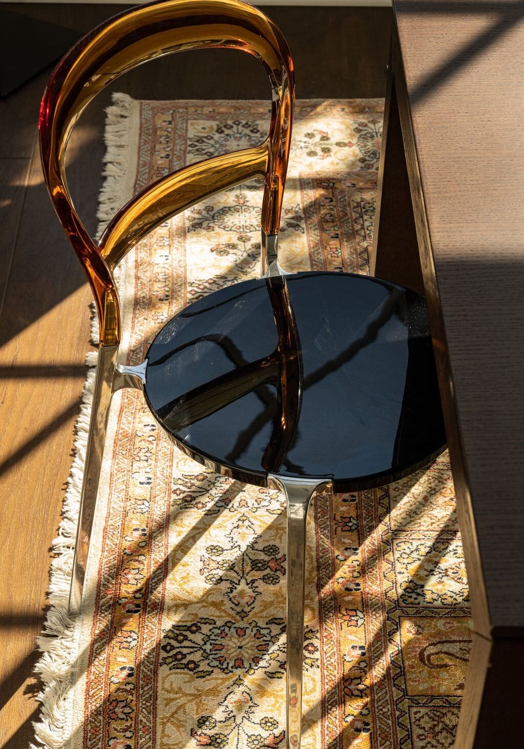 A modern chair with a translucent amber backrest and black seat is placed on a patterned rug next to a dark wooden table, with sunlight casting shadows across the scene.