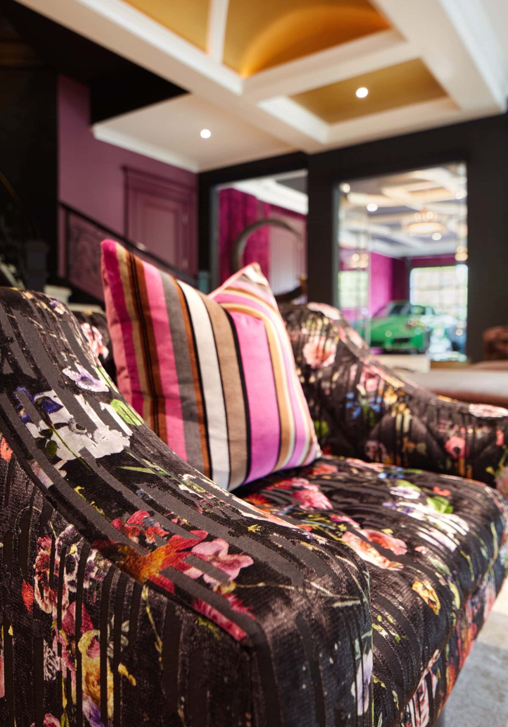 A close-up of a patterned black sofa with colorful floral designs and a striped pillow in a modern, vibrant living room with purple walls and gold ceiling accents.