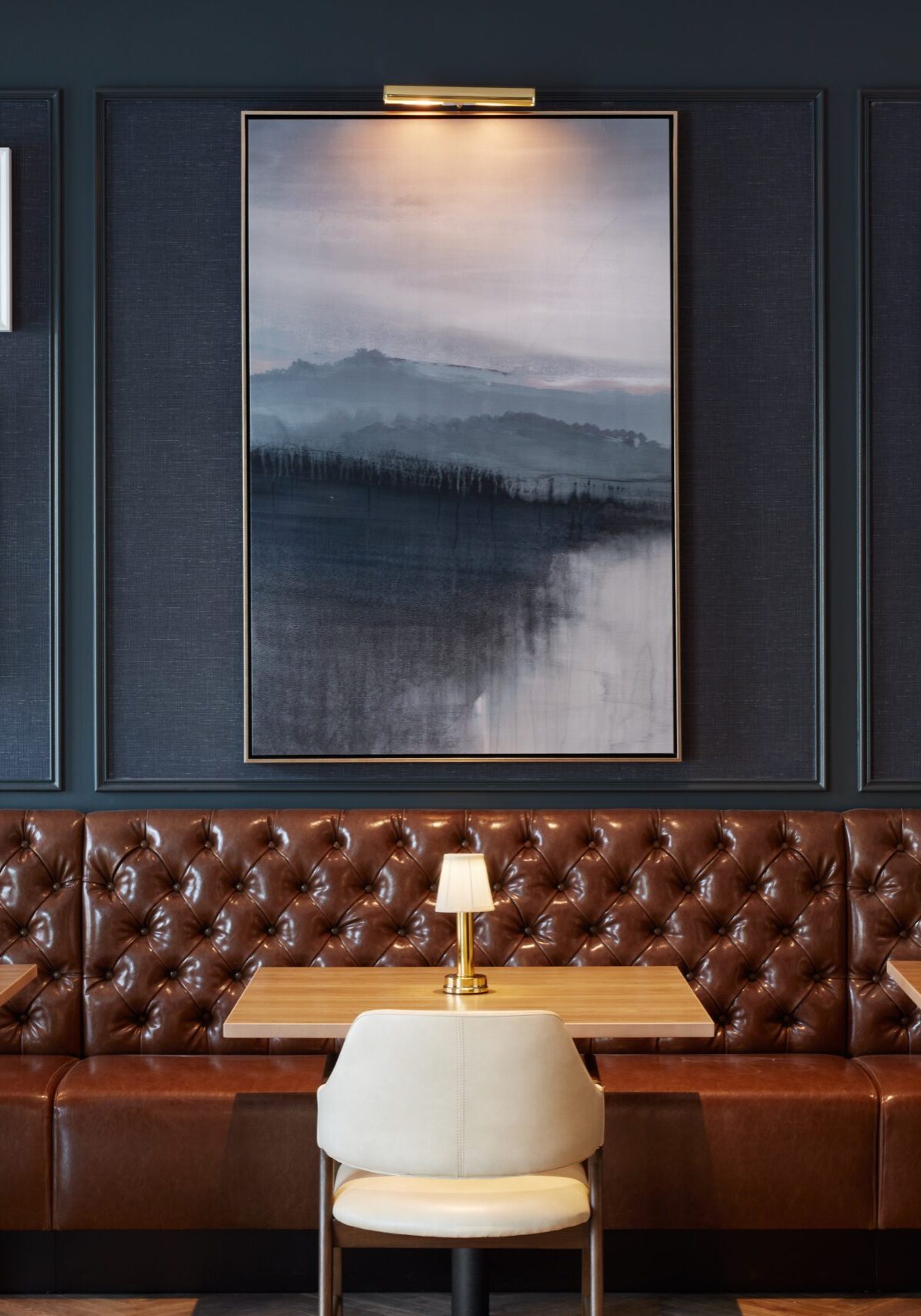 A restaurant interior with brown leather banquette seating, light wood tables, cream chairs, and framed artwork on a dark wall. Each table has a small lamp.