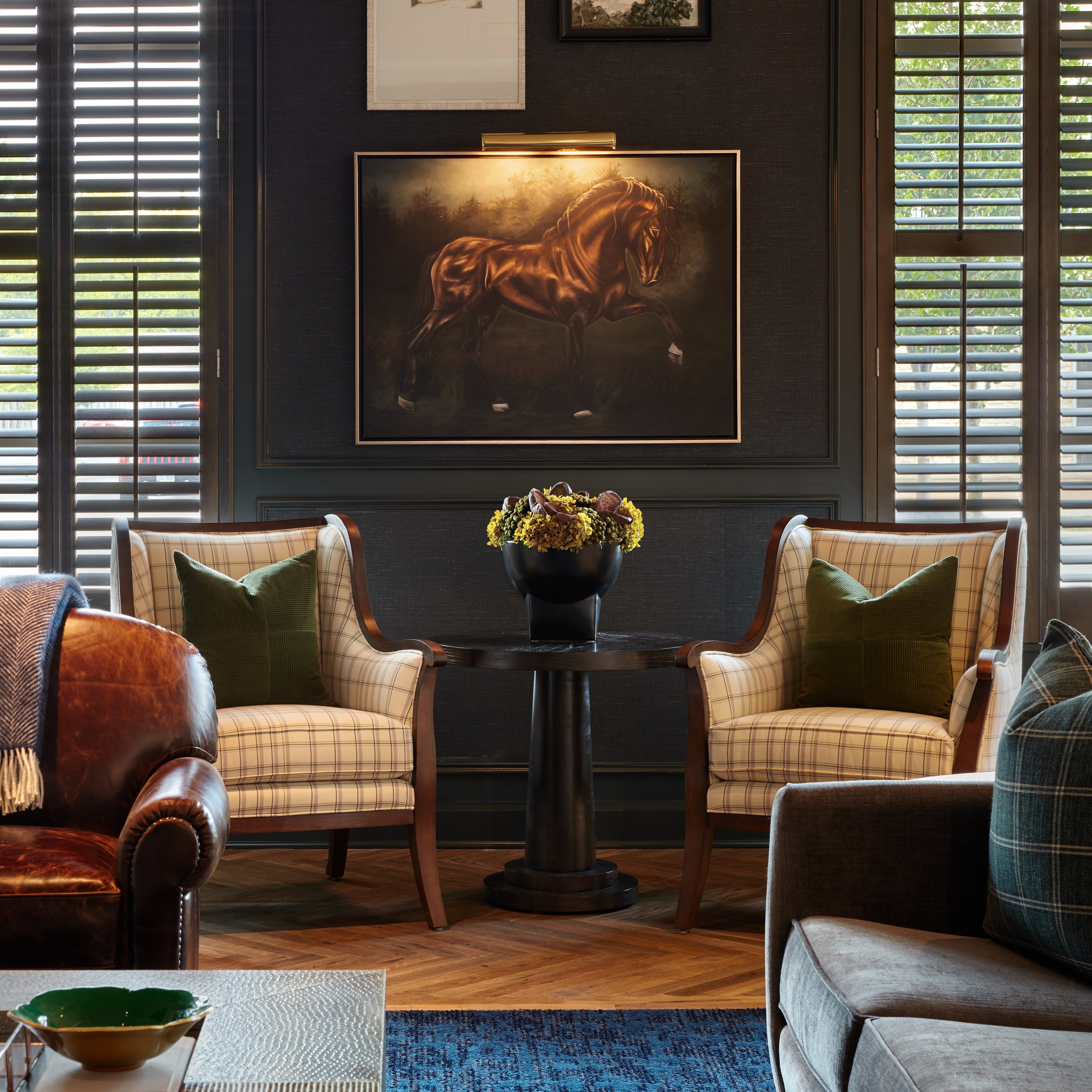 A living room by Crimson Design Group featuring two plaid armchairs, a small table with a potted plant, a horse painting, framed artwork, and large windows with shutters.