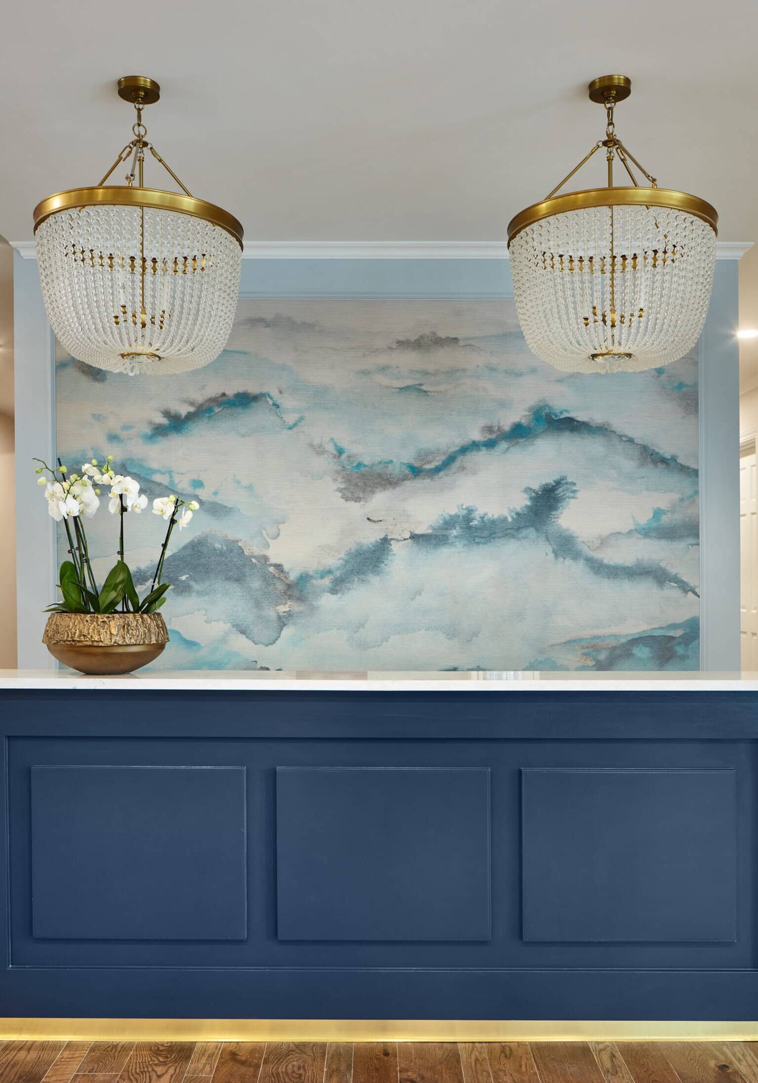 A blue reception desk with a white marble top sits on a wooden floor, in front of a blue and white abstract mural. Two gold and crystal chandeliers hang above, with an orchid on the desk.