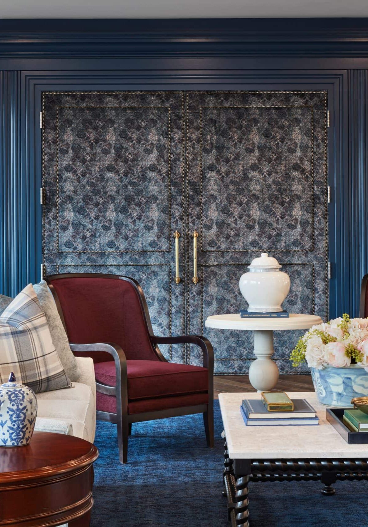 A living room with blue walls, two red armchairs, a sofa, a white coffee table with flowers, and framed landscape paintings on either side of patterned double doors.