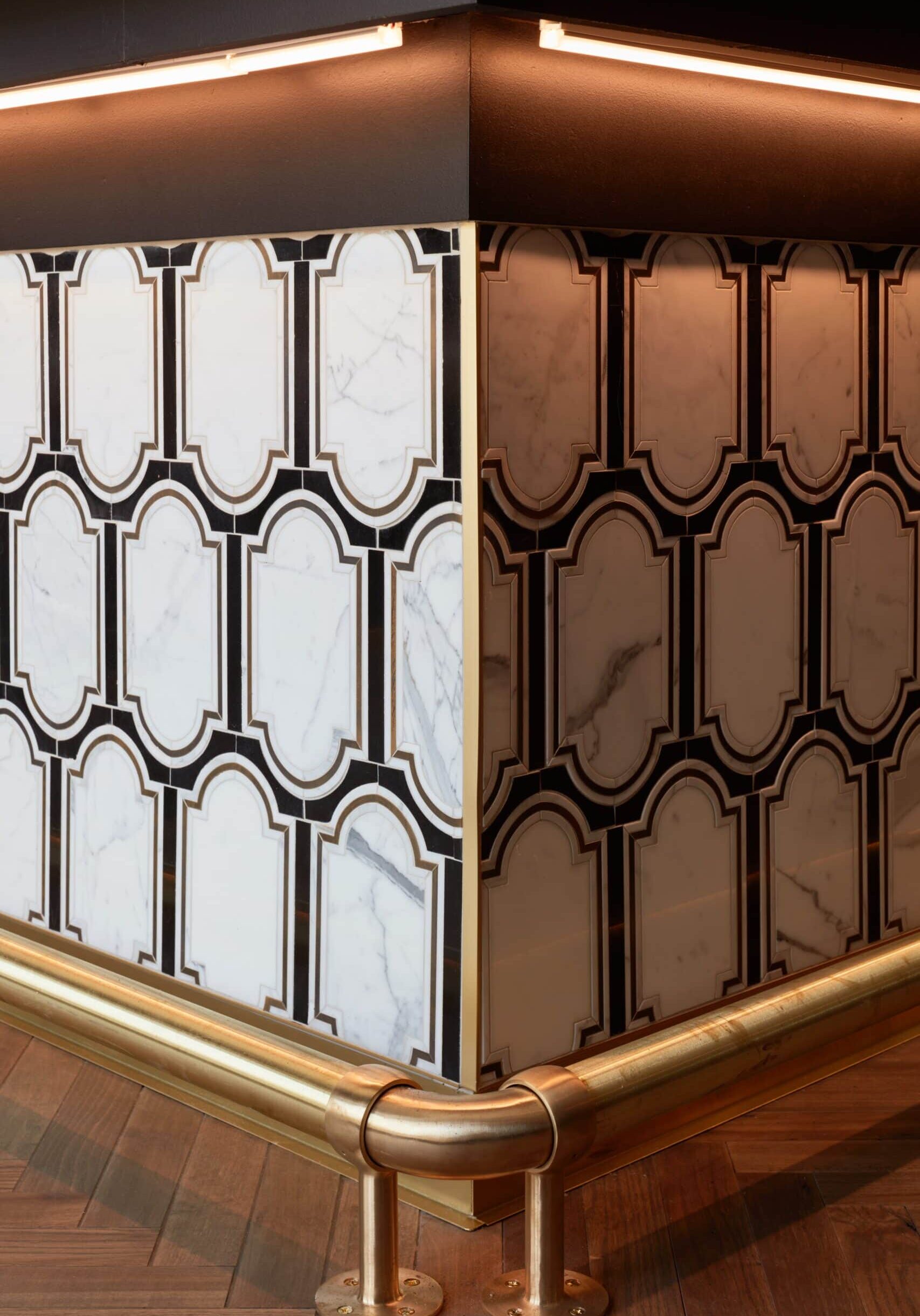Corner of a marble and brass-clad counter with geometric black, white, and gold tile pattern, wooden floor, and integrated lighting above.