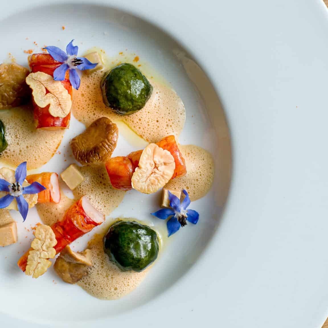 A white plate with artfully arranged gourmet food, including green spheres, diced vegetables, foam, mushrooms, edible flowers, and garnishes sits on a wooden table—an aesthetic presentation worthy of Crimson Design Group’s signature style.