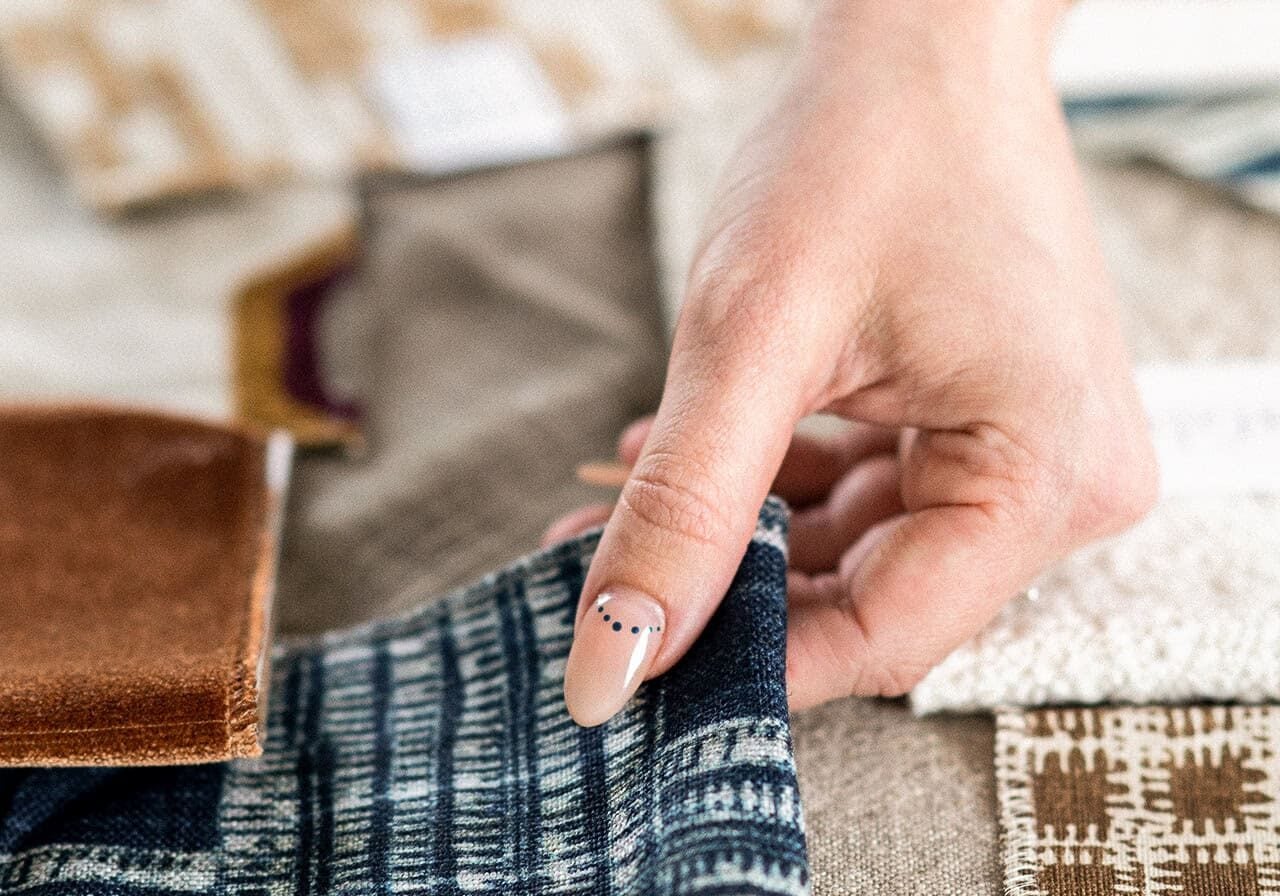 A hand touching a piece of blue patterned fabric, surrounded by various fabric swatches in different colors and textures on a table.