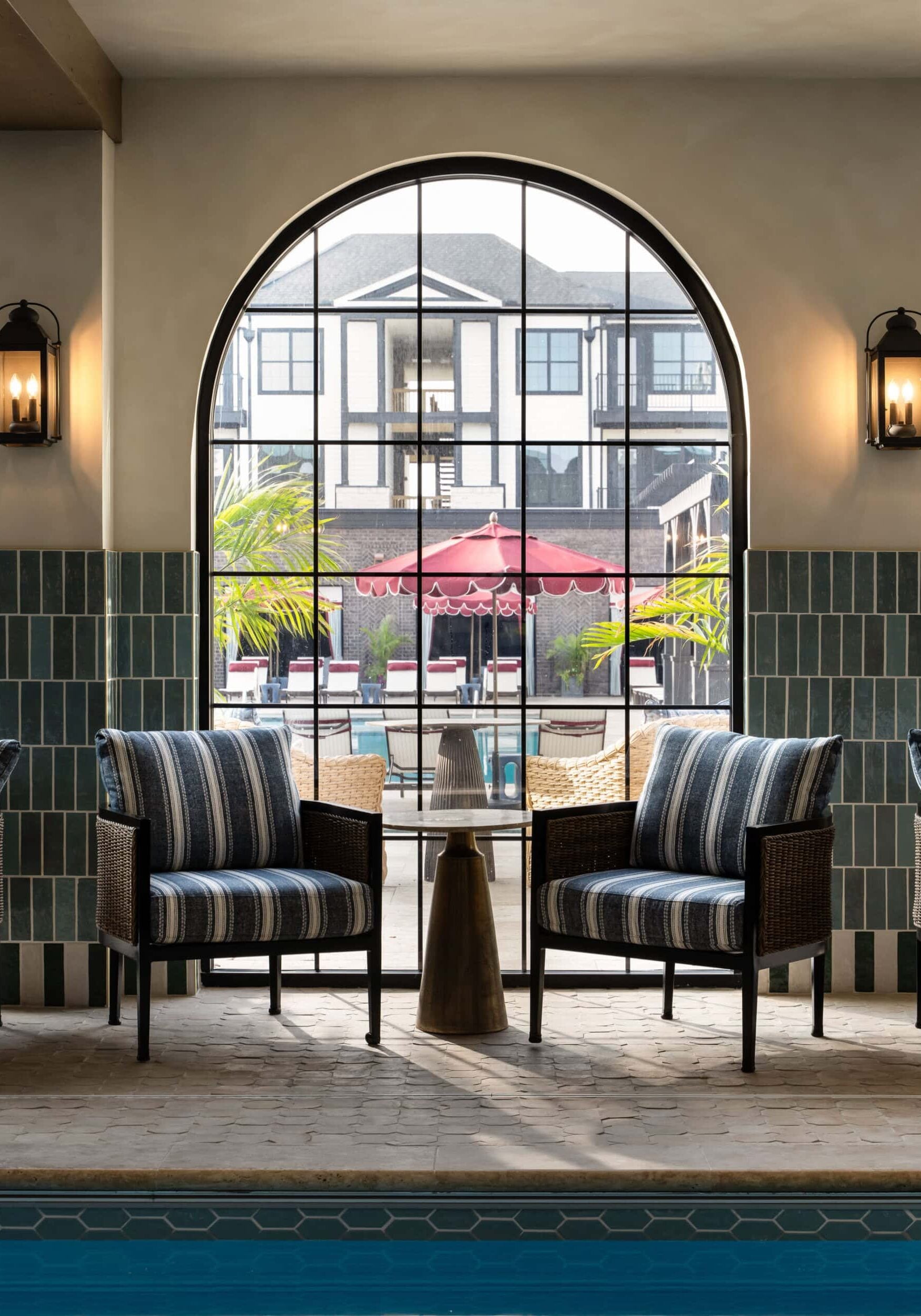 Two striped armchairs and a small table sit by an indoor pool, reflecting the elegance of retail design in Scottsdale, Arizona. A large arched window reveals an inviting outdoor patio with vibrant red umbrellas.