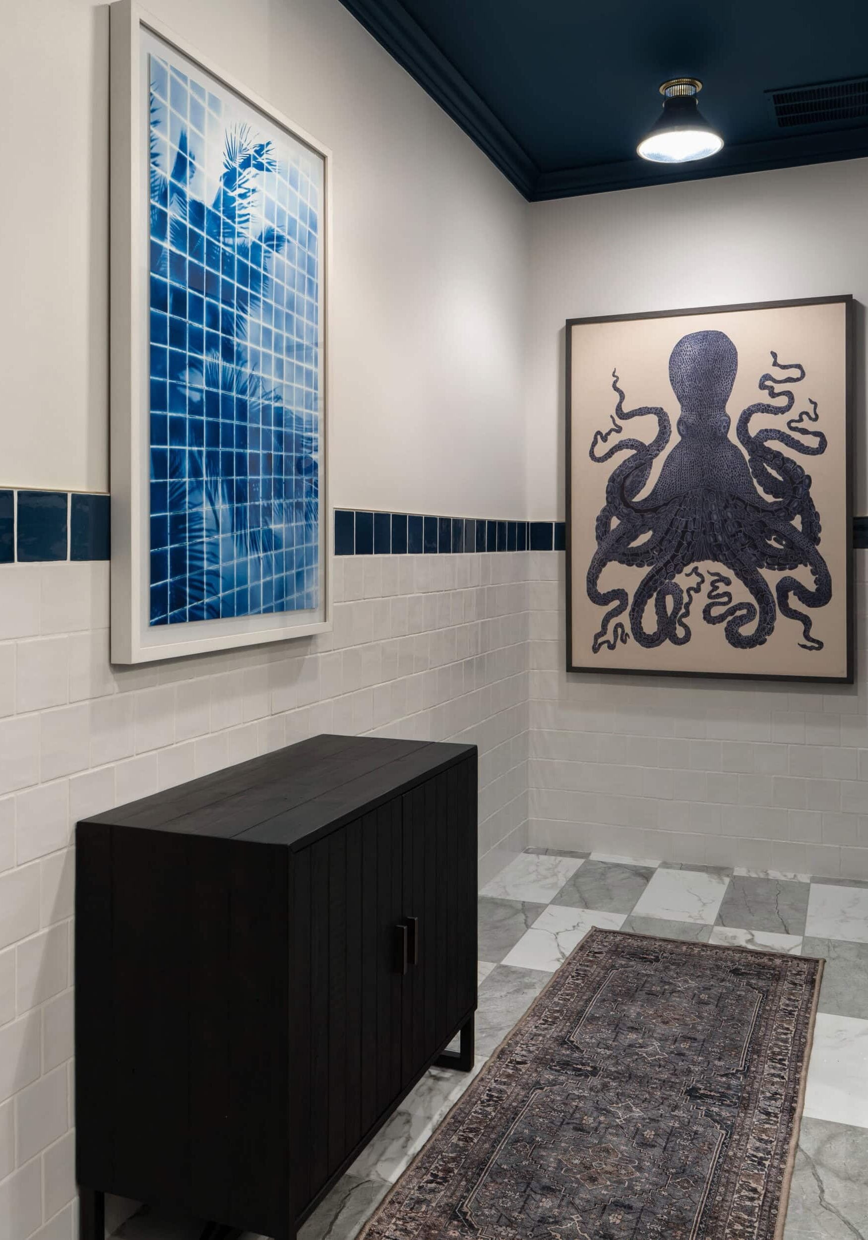Hallway with a dark cabinet, patterned rug, blue tile accents, and two framed artworks: one of a blue grid with leaves and another depicting a dark octopus. Marble floor and overhead light create an inviting space reminiscent of contemporary retail design in Scottsdale, Arizona.
