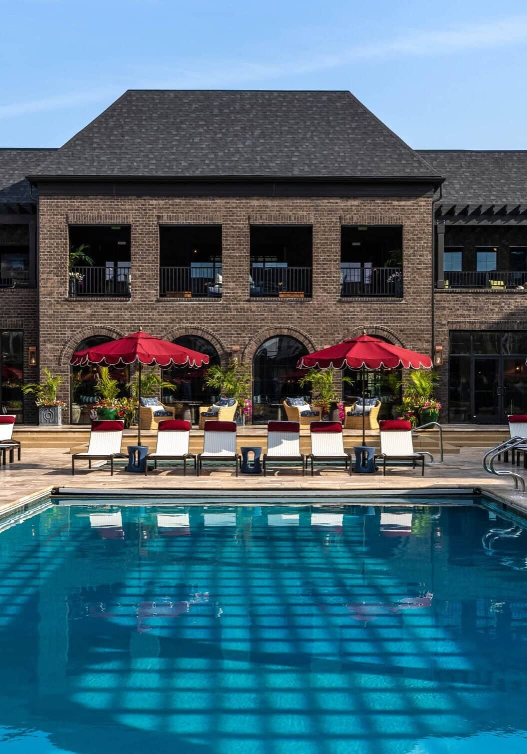 A large outdoor pool with lounge chairs and red umbrellas, set in front of a two-story brick building under a blue sky, showcases the sophisticated flair of retail design in Scottsdale, Arizona.