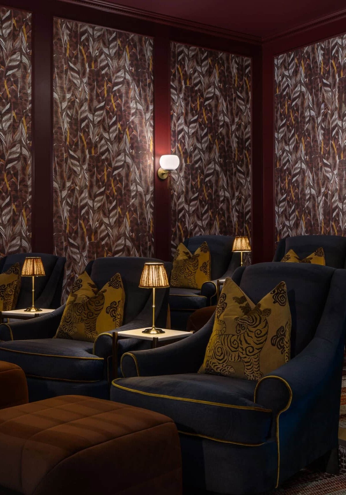A dimly lit room with dark armchairs, patterned pillows, small lamps, ottomans, and feather-patterned wallpaper—expertly arranged for cozy seating—reflects the sophisticated touch of retail design in Scottsdale, Arizona.