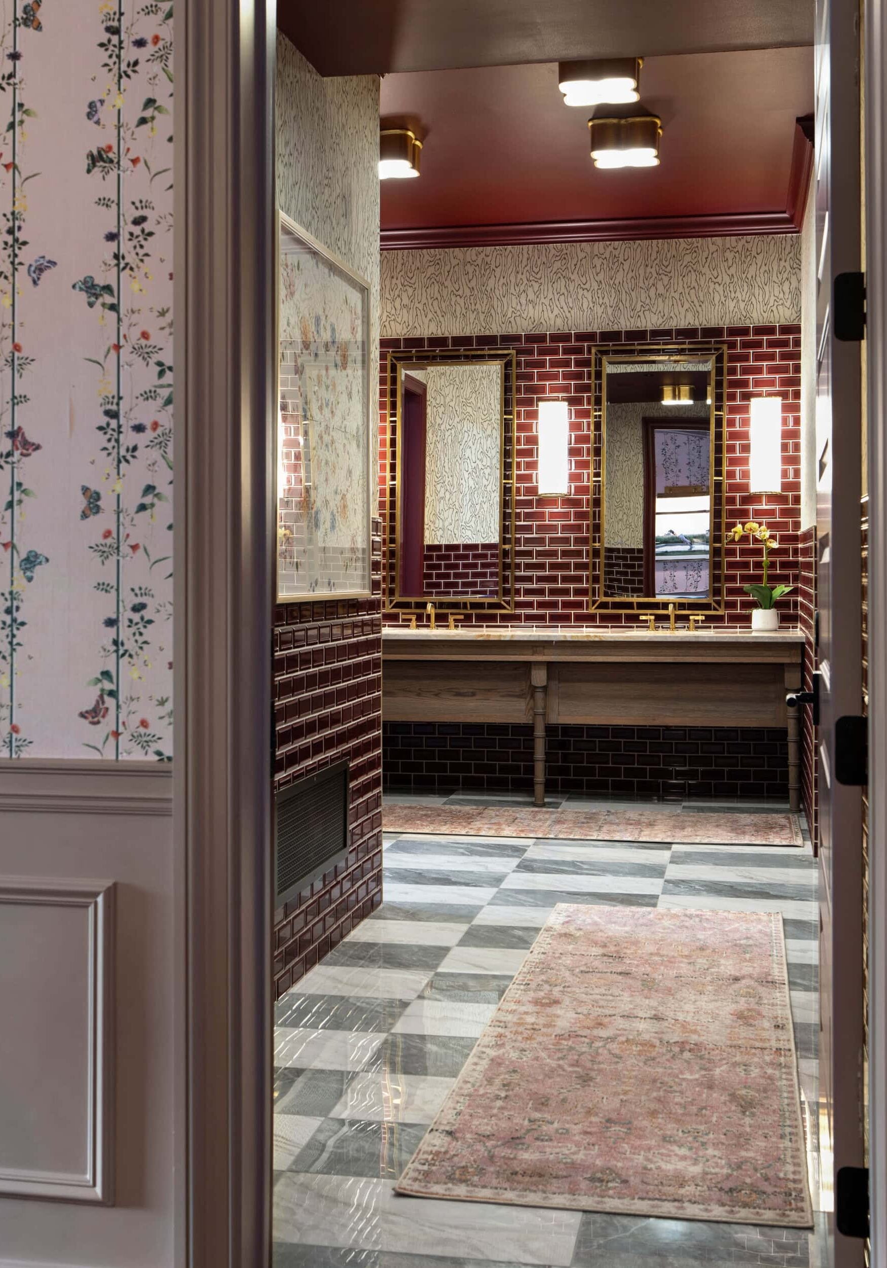 A view into a bathroom with brick walls, patterned wallpaper, three mirrors, a double sink, plaid flooring, and a pink patterned rug—showcasing the unique flair of retail design in Scottsdale, Arizona.