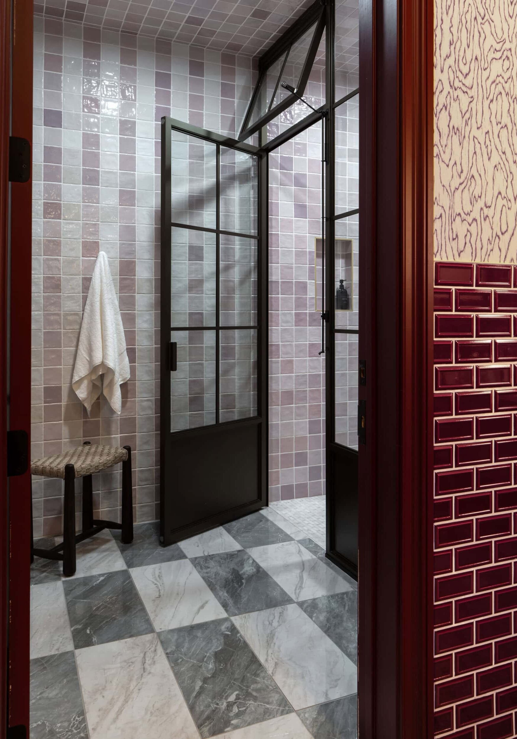 Bathroom with checkered gray marble floor, pink and white tiled shower, glass door, towel on a stool, and maroon tile accents—showcasing a refined aesthetic inspired by retail design in Scottsdale Arizona.