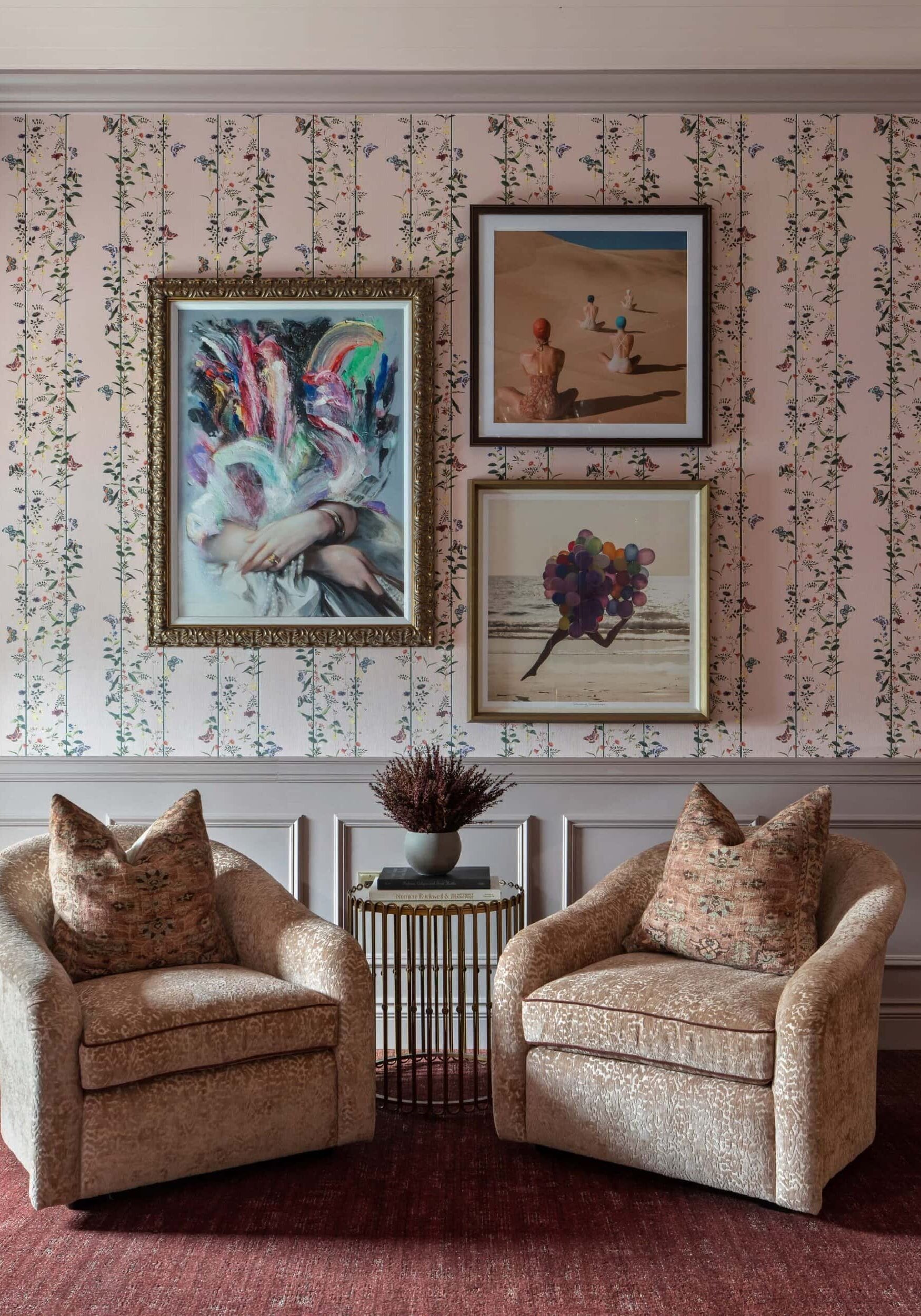 Two beige armchairs with patterned pillows flank a small round table with a plant. Behind, floral wallpaper is decorated with four framed art pieces arranged in a grid, showcasing an elegant touch inspired by retail design in Scottsdale, Arizona.