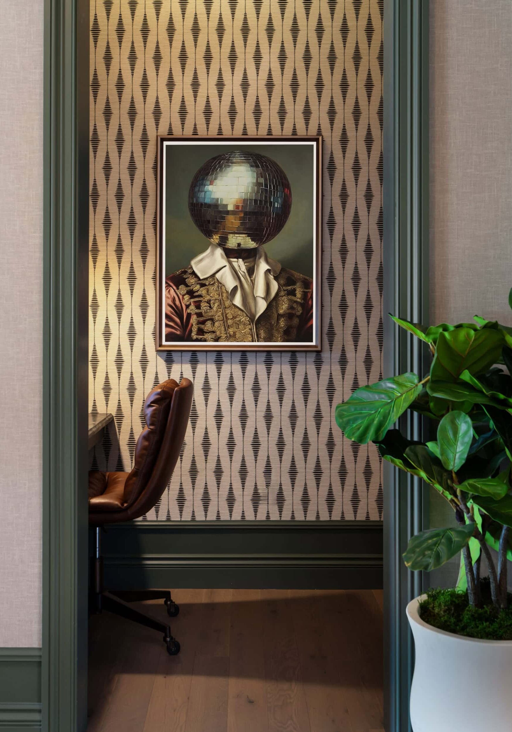 A framed painting of a figure with a disco ball for a head hangs on a patterned wall in a small alcove, next to a desk, chair, and potted plant—an artful vignette that captures the innovative spirit often seen in Retail Design in Scottsdale, Arizona.