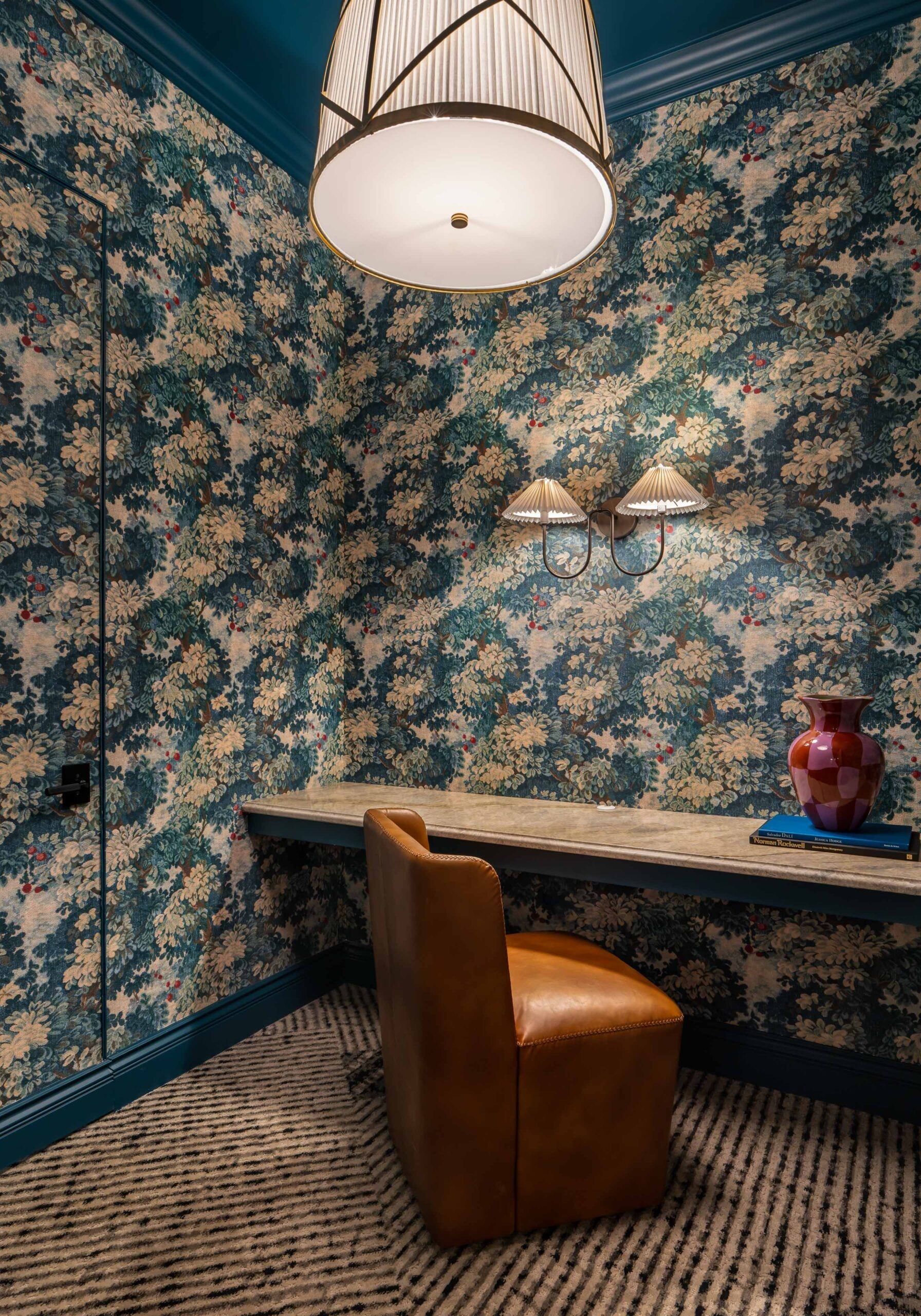 A small room, inspired by retail design in Scottsdale, Arizona, features blue floral wallpaper, a built-in desk, a tan leather chair, a vase, two wall sconces, and a ceiling pendant light over striped carpet.