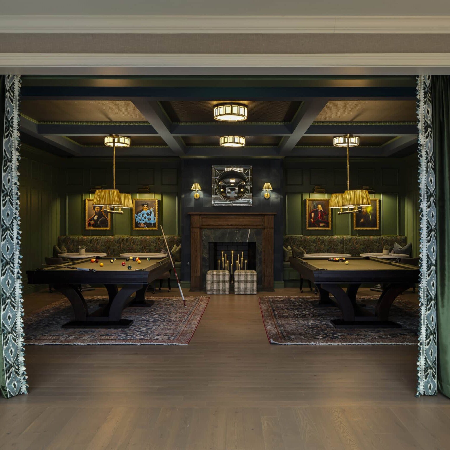 Room with green walls, two pool tables on patterned rugs, green velvet drapes, a central fireplace, and framed art on the walls. Overhead lights and lamps provide illumination.