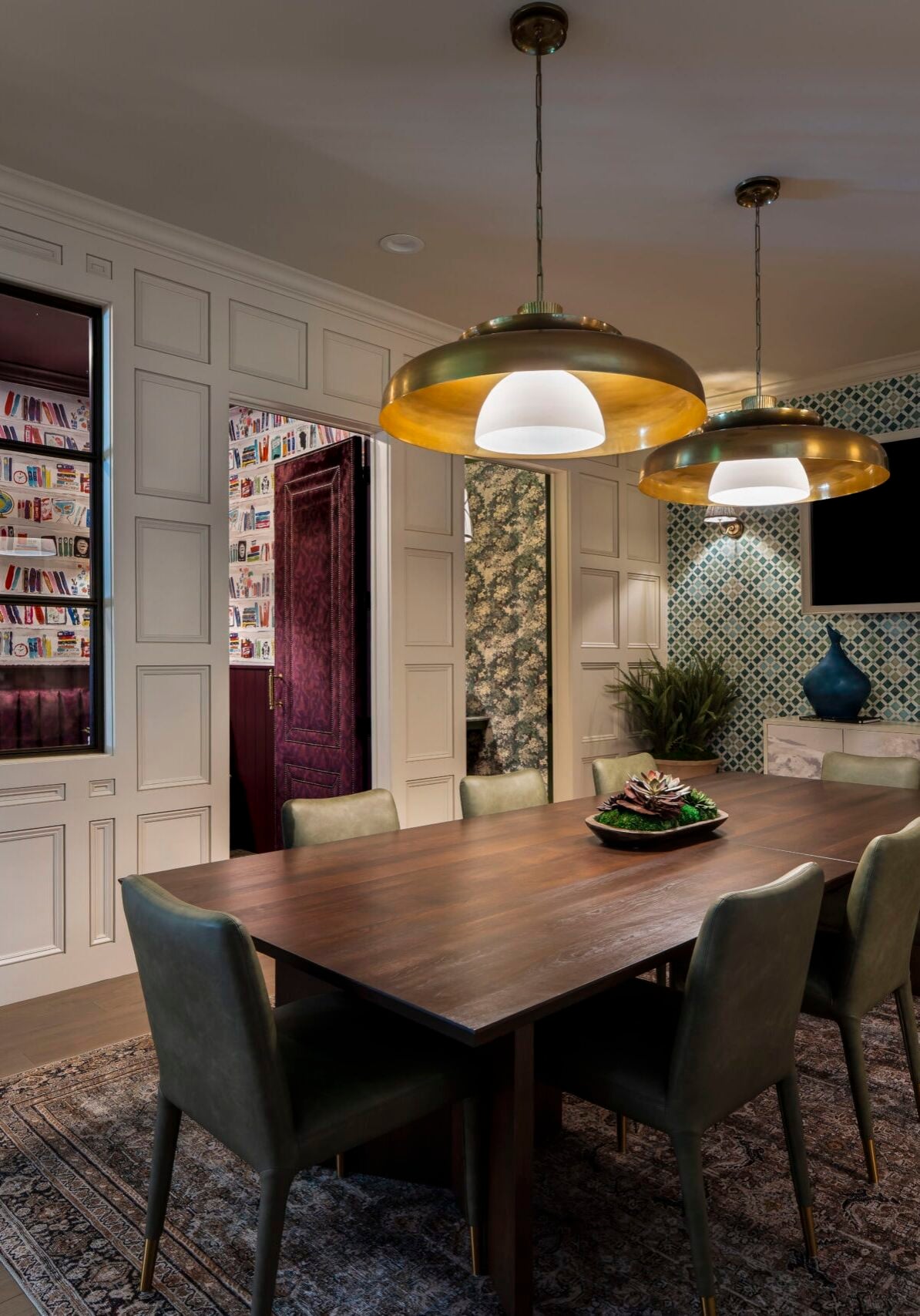 Modern dining room with a wooden table, six green chairs, three pendant lights, a wall-mounted TV, patterned wallpaper, and a window showing a colorful library.