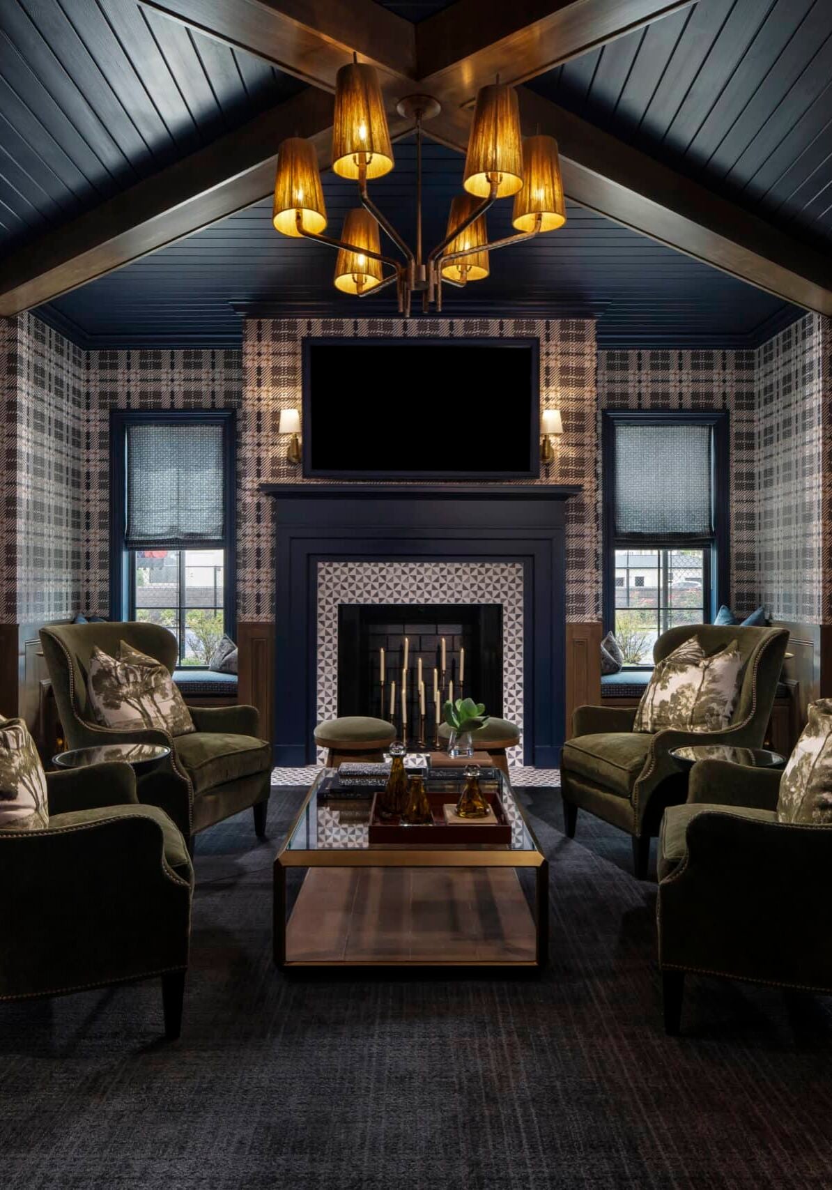 Plaid-walled lounge with six green armchairs around a wooden coffee table, fireplace, TV, chandelier, framed art, and wood-paneled door.