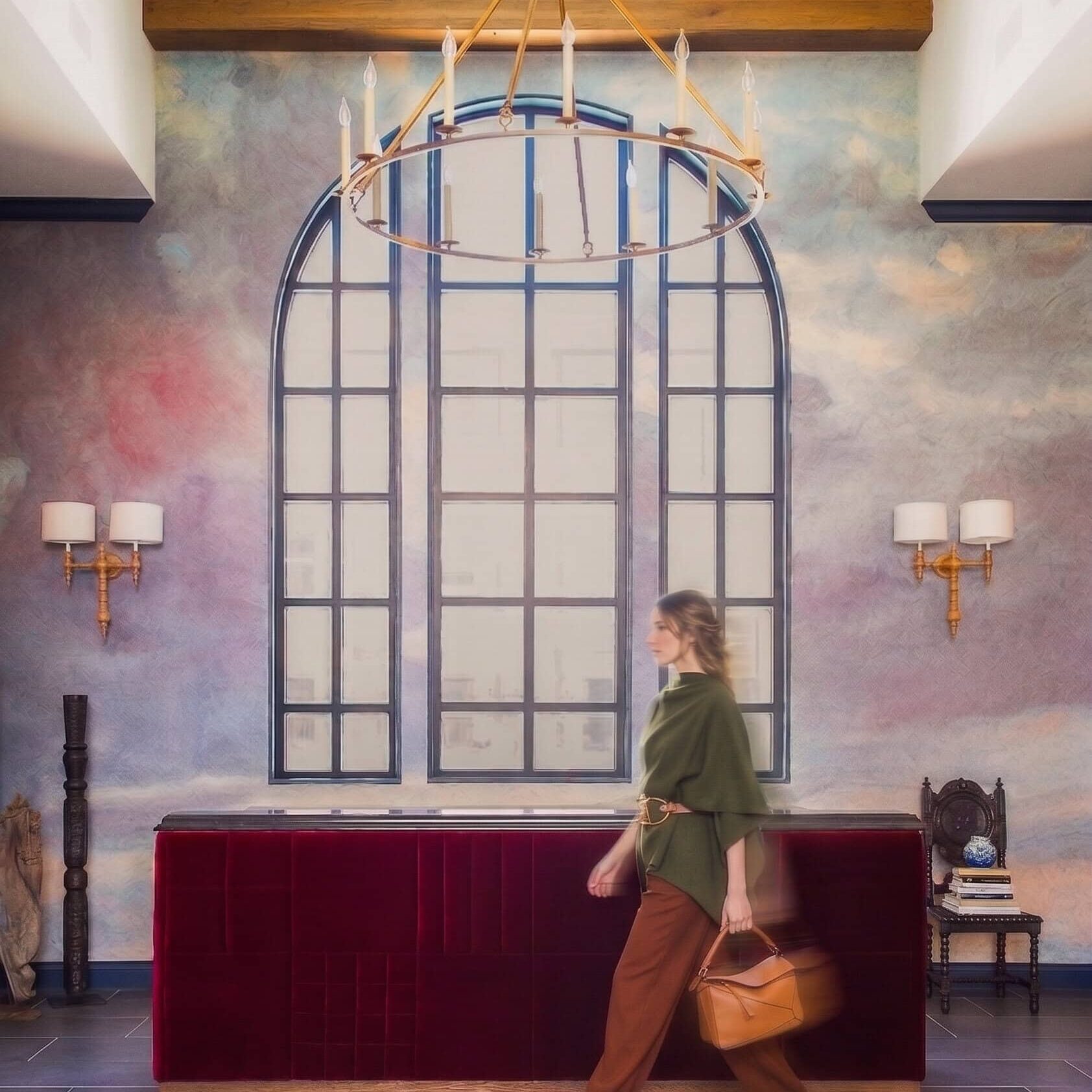 A person carrying a tan bag walks past a red reception desk in a room with a large arched window, chandelier, and decorative wall sconces.
