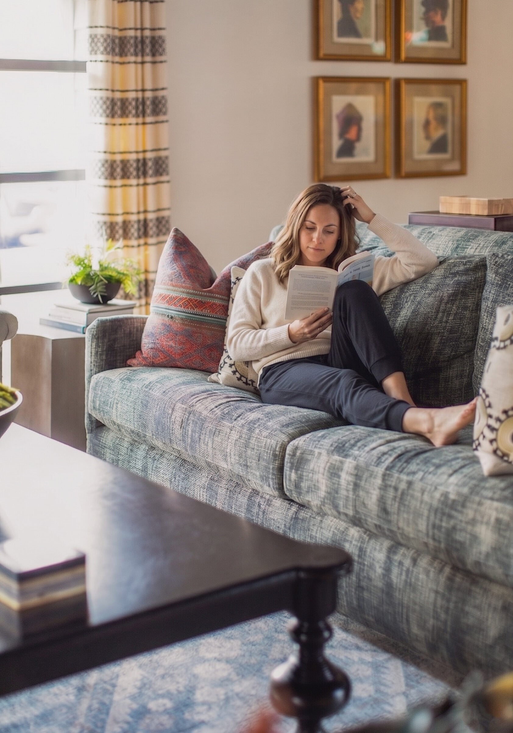 A woman sits barefoot on a sofa, reading a book in a cozy, sunlit living room with cushions, a patterned rug, and framed portraits on the wall.