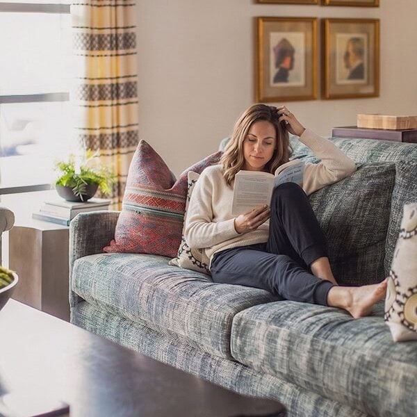 A woman sits on a blue sofa reading a book in a well-lit living room with patterned pillows, framed art, and a large window.
