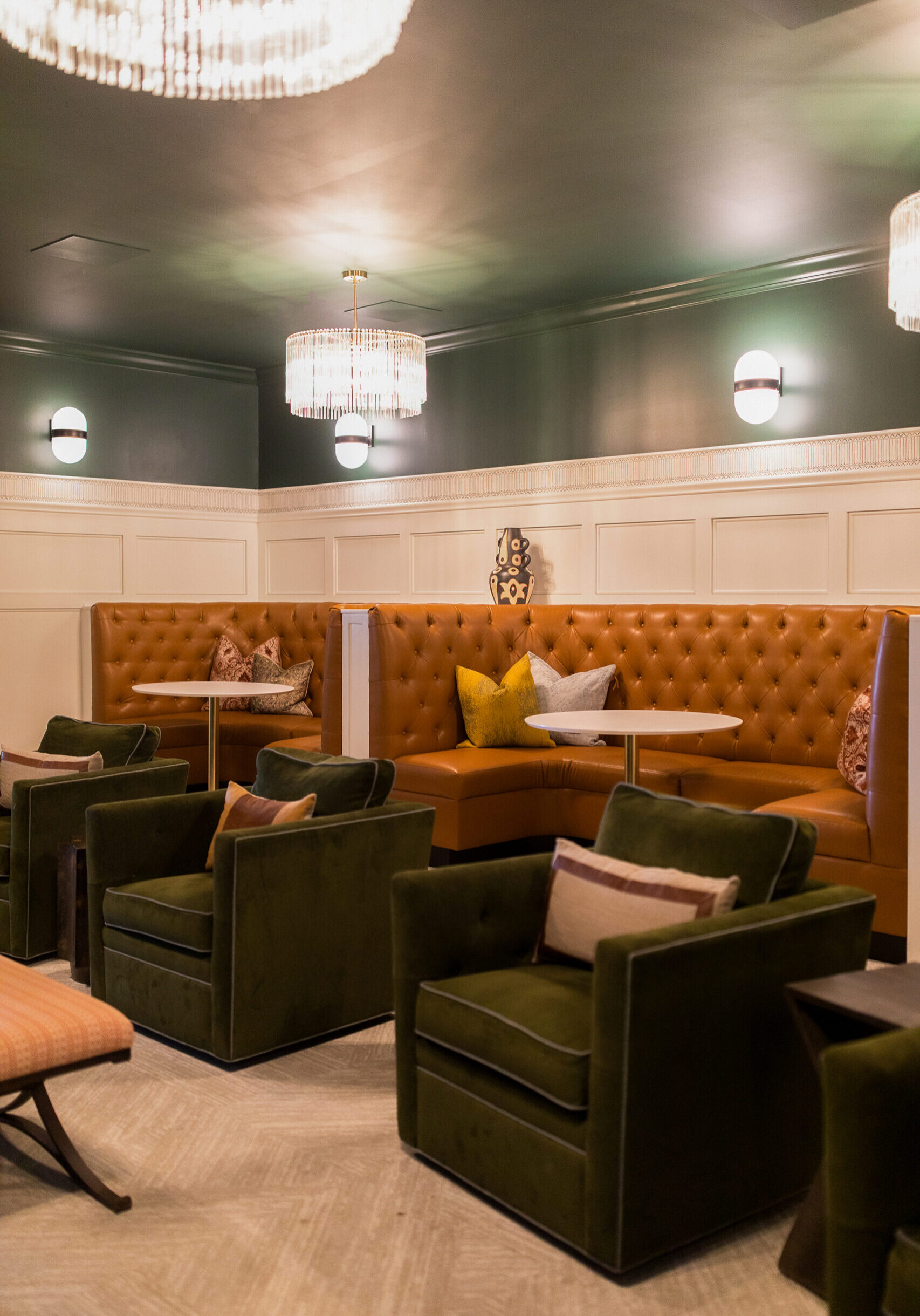 A lounge area with olive green armchairs, tan tufted banquette seating, pillows, small tables, and two ceiling chandeliers.