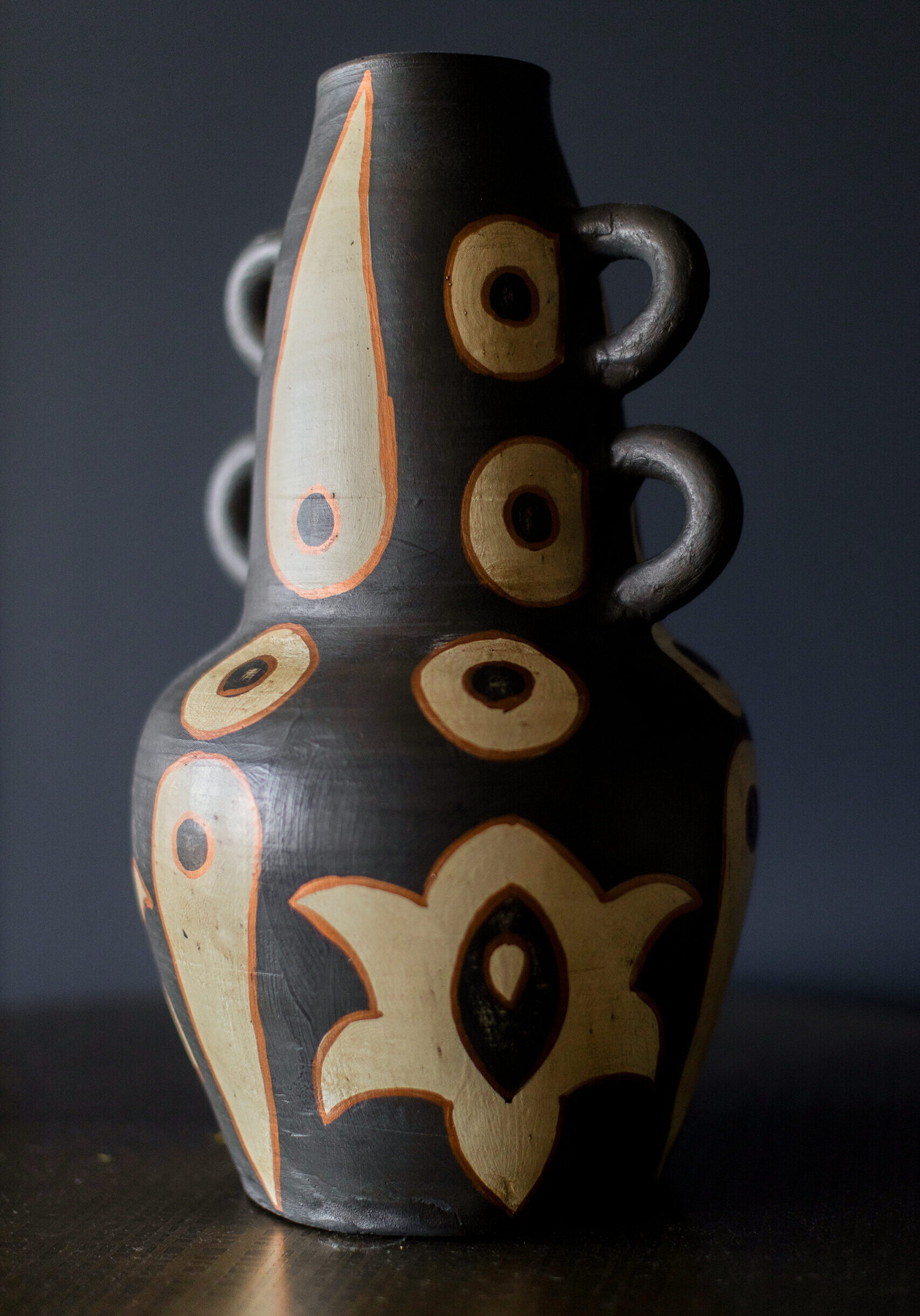 A ceramic vase with a dark matte finish and geometric beige and orange patterns, featuring three ring-shaped handles on each side, standing on a dark surface.