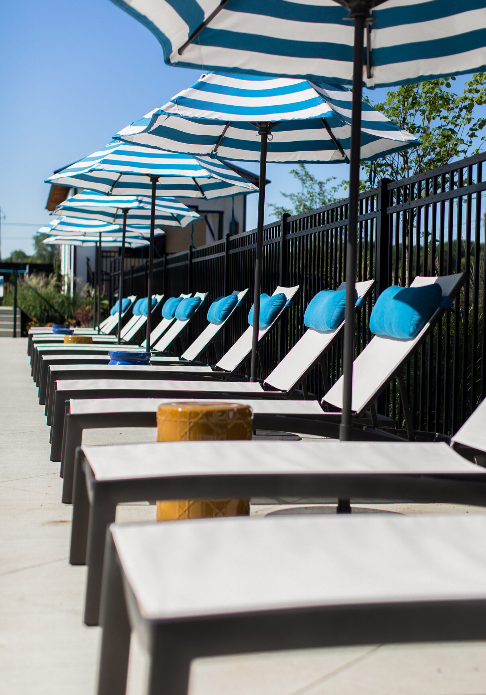 A row of empty lounge chairs with blue cushions and striped umbrellas beside a black metal fence on a sunny day.