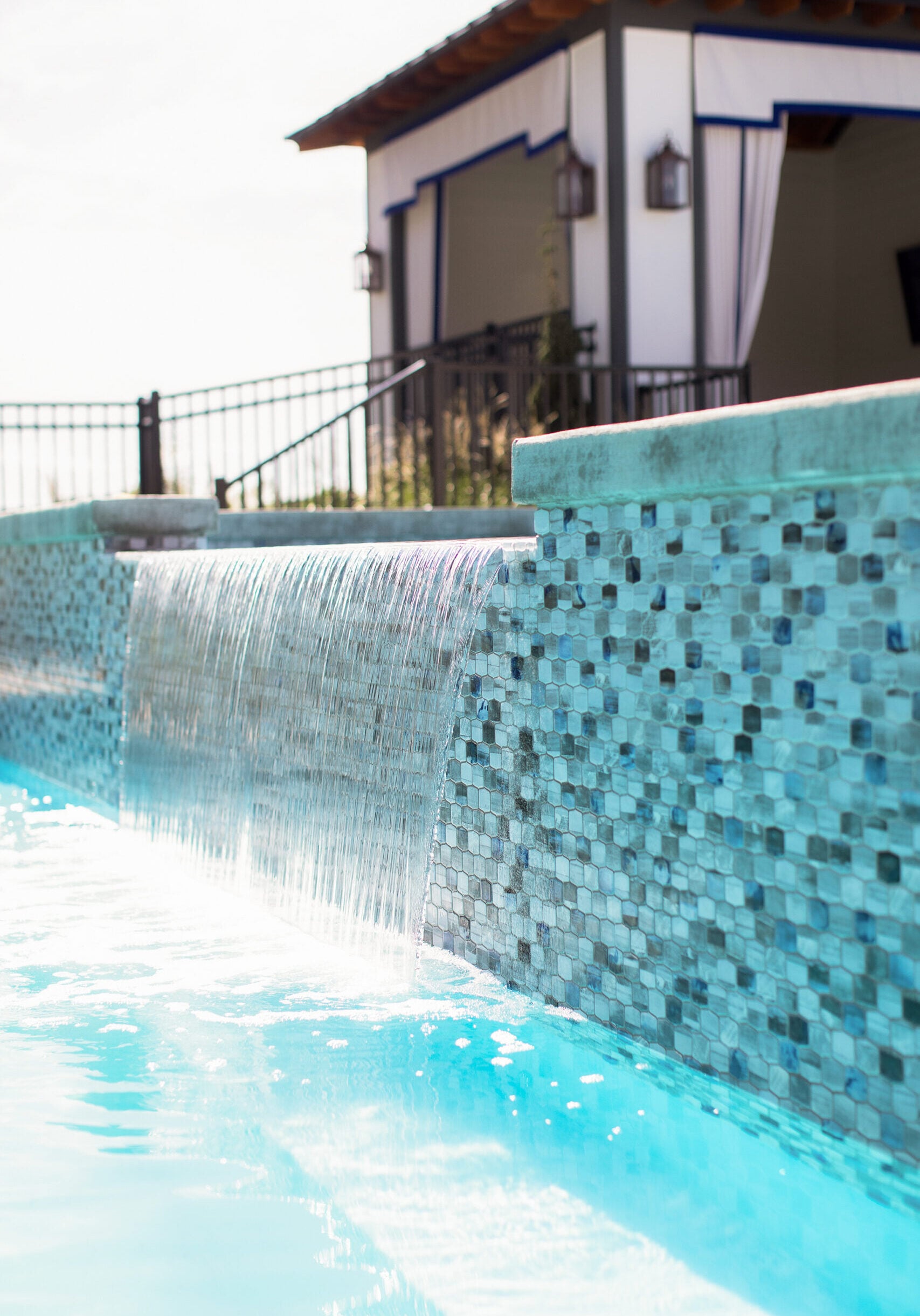 A close-up of a swimming pool with blue mosaic tiles, featuring a waterfall edge and a poolside cabana in the background.