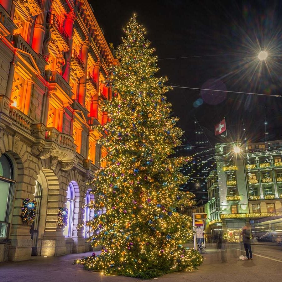 A large Christmas tree decorated with lights stands on a city street at night, its festive glow echoing the elegance of a Crimson Design Group creation, with illuminated buildings and passing pedestrians nearby.