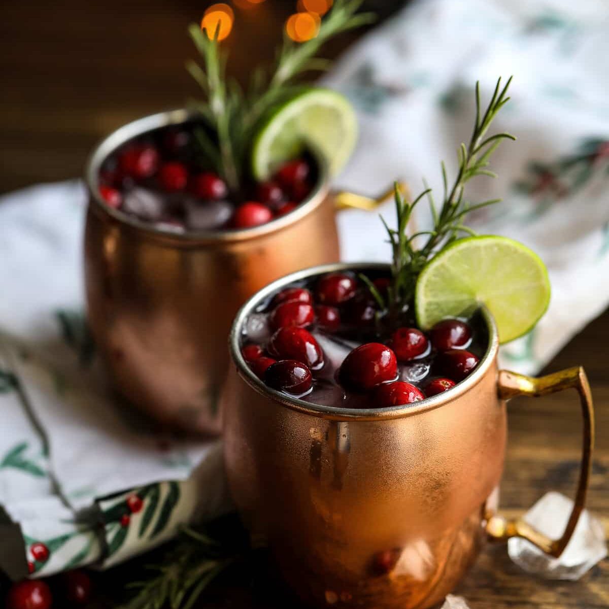 Two copper mugs filled with a dark red drink, garnished with cranberries, lime slices, and rosemary, sit on a wooden table with ice cubes and a festive cloth—a cozy scene inspired by Crimson Design Group.