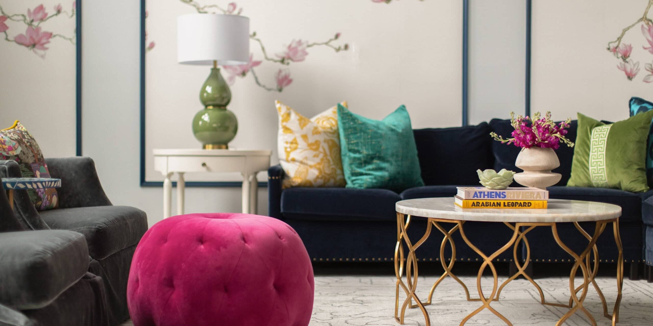 A living room with a dark sofa, colorful pillows, a round marble coffee table, a pink tufted ottoman, and floral wallpaper in the background.