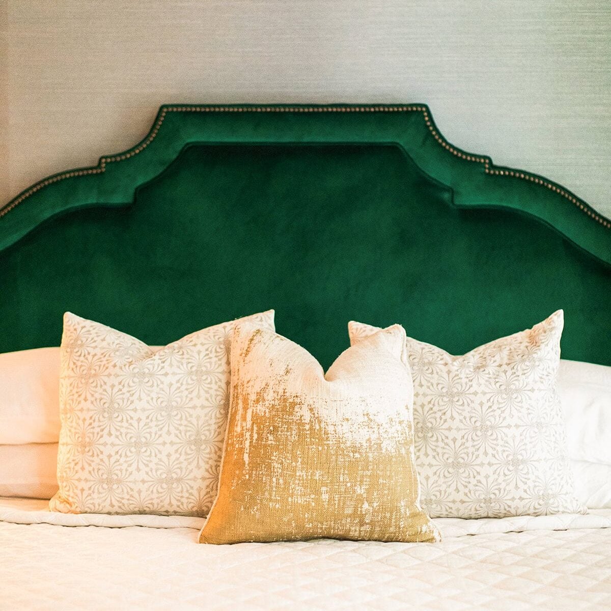 A neatly made bed with a green upholstered headboard, two white patterned pillows, and a gold decorative pillow in the center.