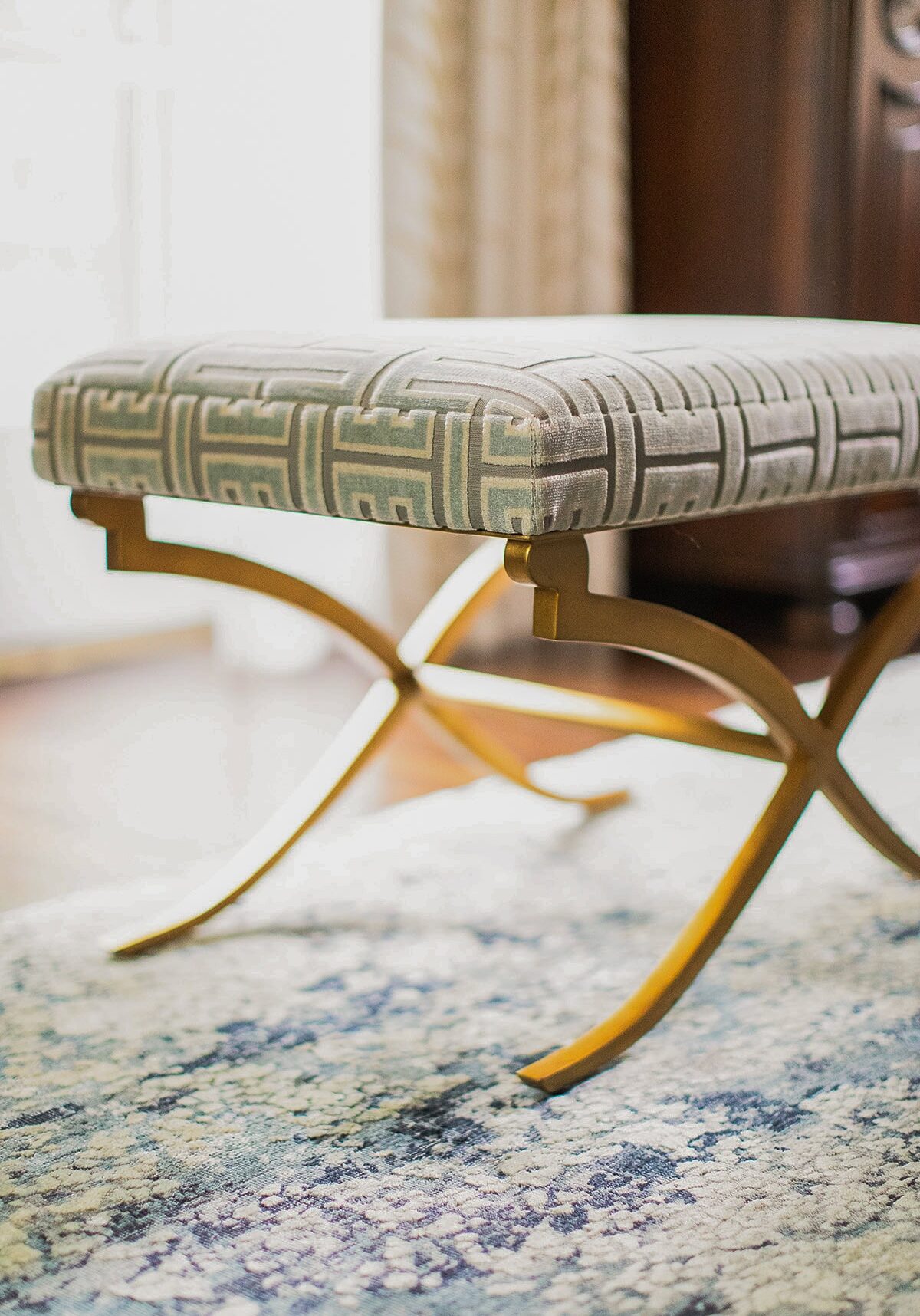Upholstered bench with geometric patterned fabric and gold metal legs on a blue-patterned rug in a softly lit room.