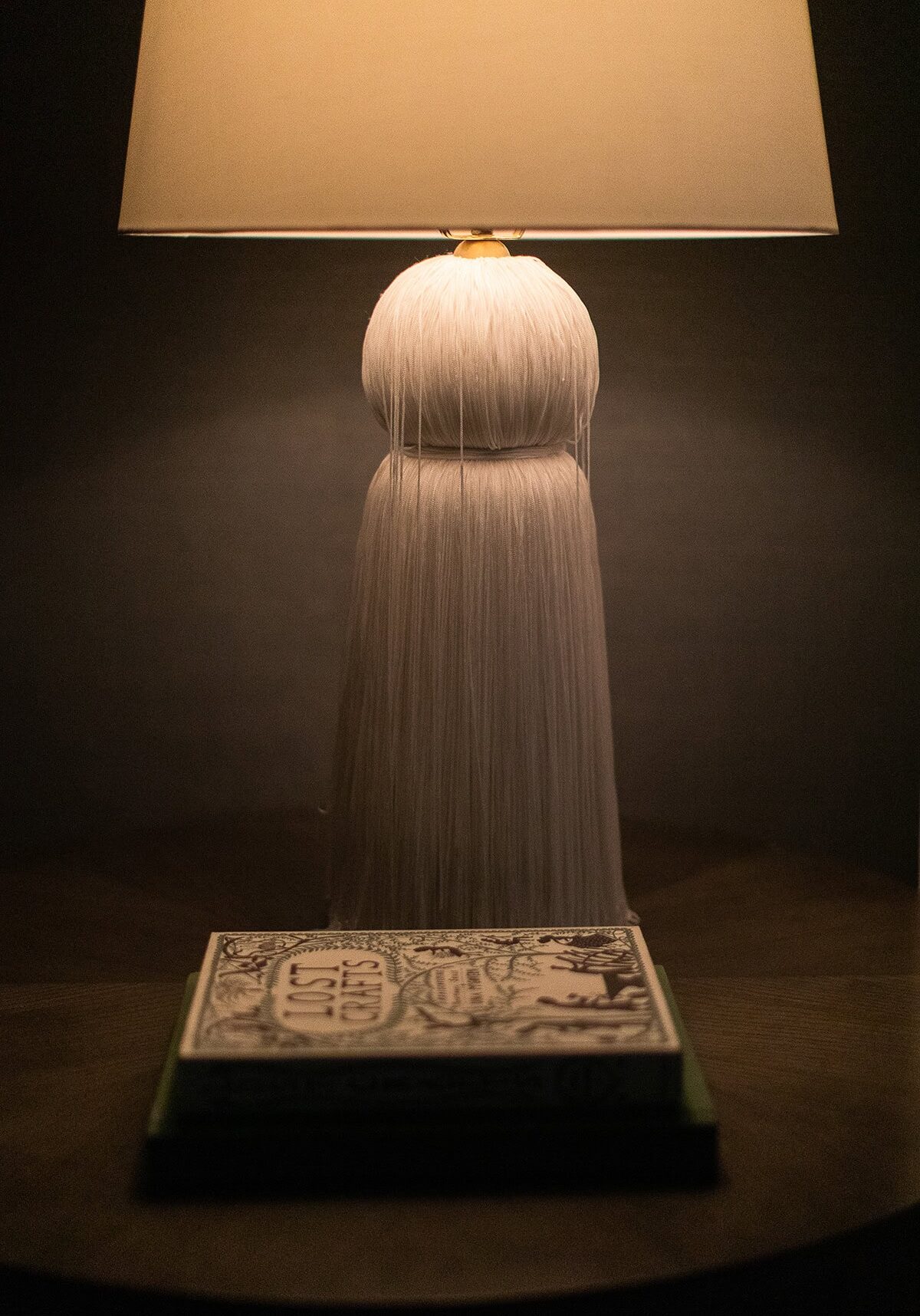 A table lamp with a textured white base and a beige shade sits on a wooden surface beside two stacked books, one titled "Alice's Adventures in Wonderland.