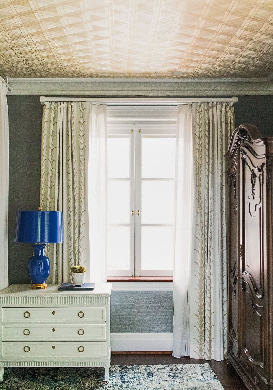 A window with white curtains is centered between a white nightstand with a blue lamp and a dark wooden wardrobe in a bedroom.